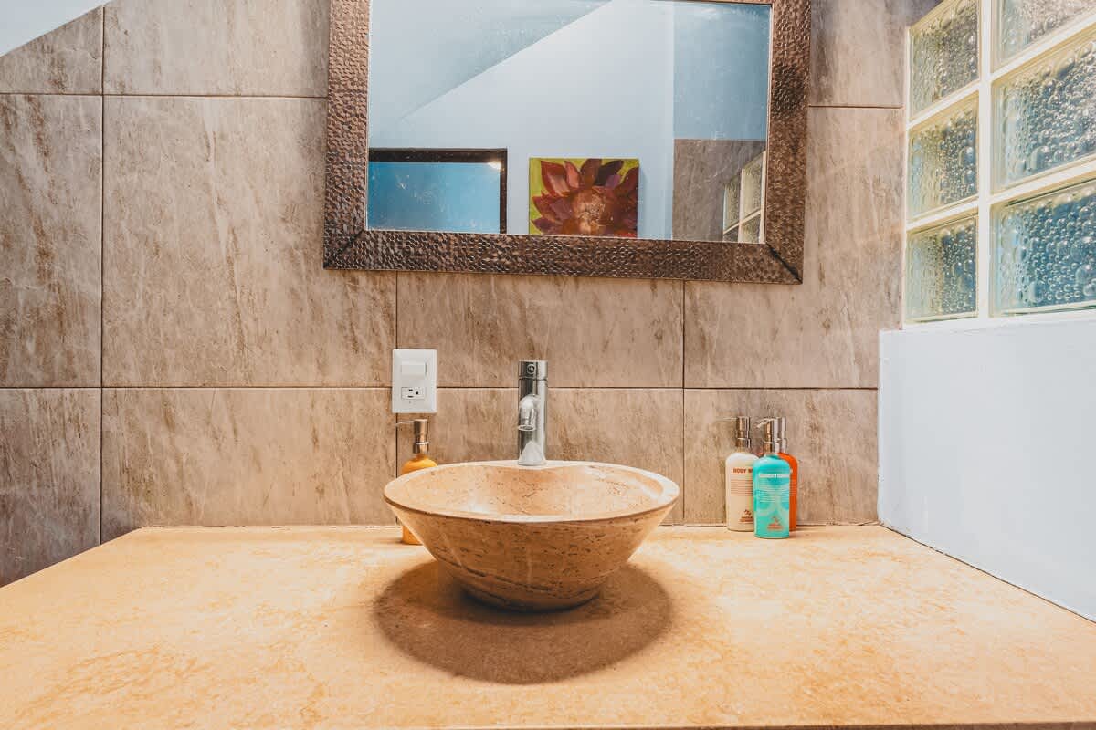 A bathroom vanity area with light beige marble-look tiles, a unique stone vessel sink, and a rustic framed mirror above. Various toiletries are neatly arranged on the countertop, complemented by glass block windows on the right.