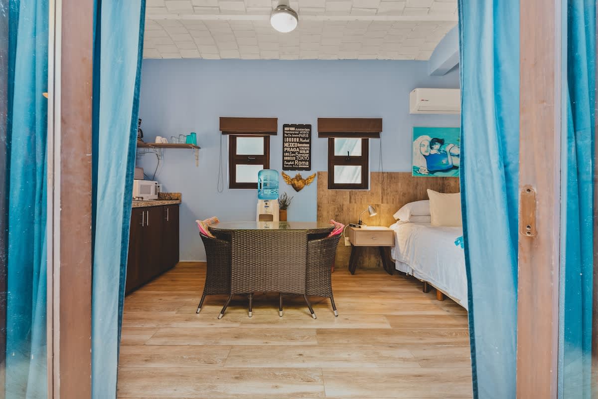A studio apartment entrance opening into a combined kitchen and dining area, featuring a glass-top table with wicker chairs, a small kitchen counter, and a bed visible further inside, all with light blue walls and wood-look flooring.