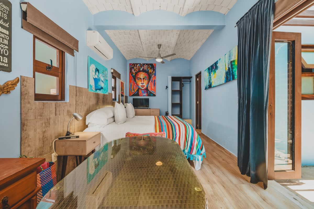 A colorful studio bedroom with light blue walls and a vaulted brick ceiling, featuring a large bed with a striped blanket and a glass-top table in the foreground. Vibrant artwork, including a large portrait, decorates the walls, and a dark curtain covers a doorway.