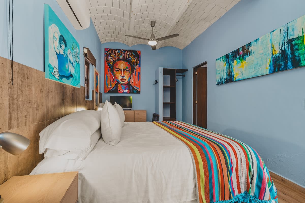 A vibrant bedroom with light blue walls, a white vaulted brick ceiling, and a large bed adorned with a colorful striped blanket. Artwork, including a prominent portrait, decorates the walls, and a ceiling fan hangs above.