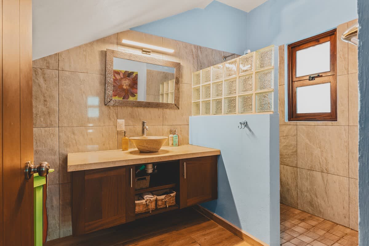 A spacious bathroom with light beige tiled walls and a light blue accent wall, featuring a floating dark wood vanity with a stone vessel sink and a large framed mirror. Glass block windows provide separation to the tiled shower area.