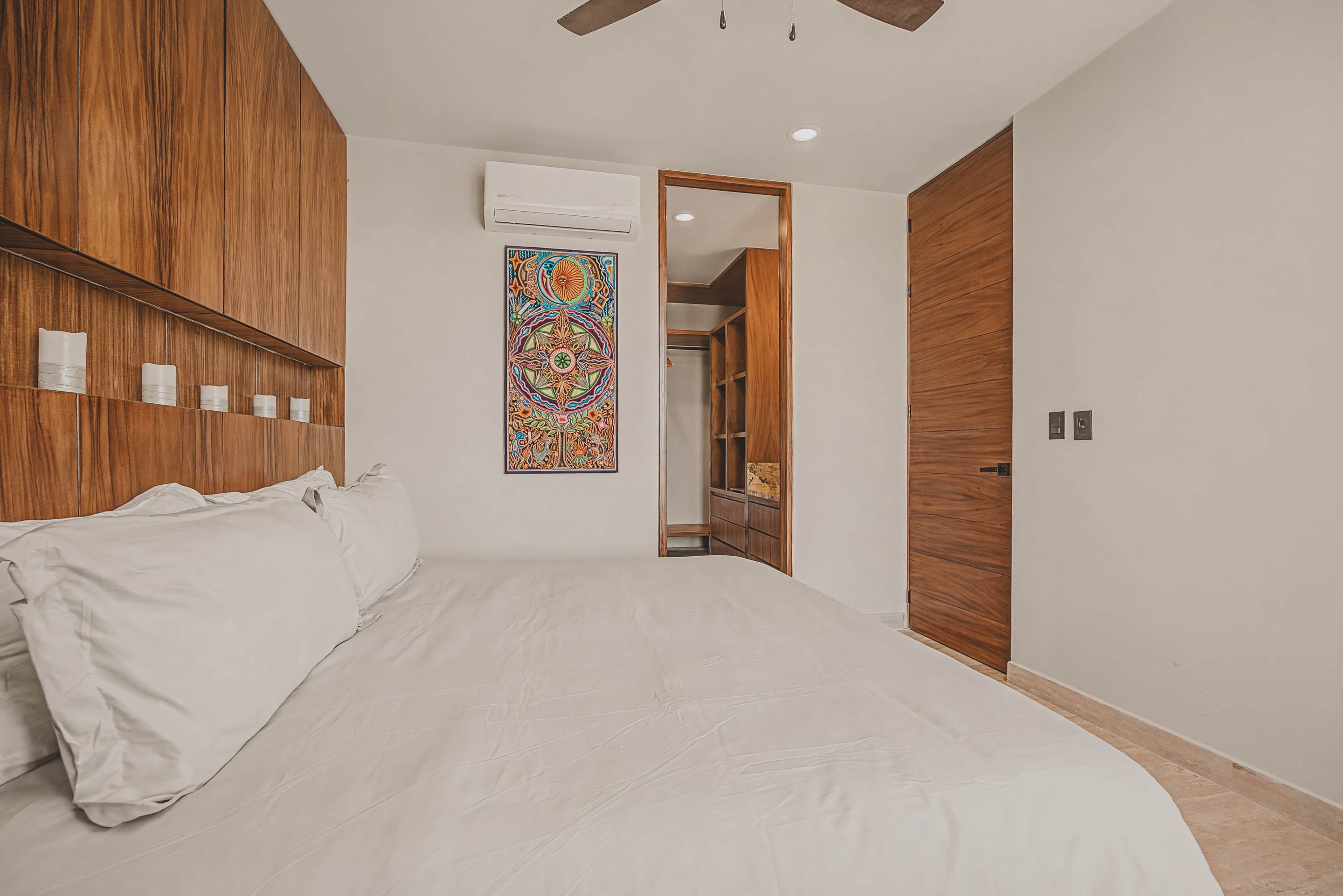 Modern bedroom with a white bed, a striking wooden panel headboard, and an open doorway leading to a walk-in closet with built-in wooden shelving.