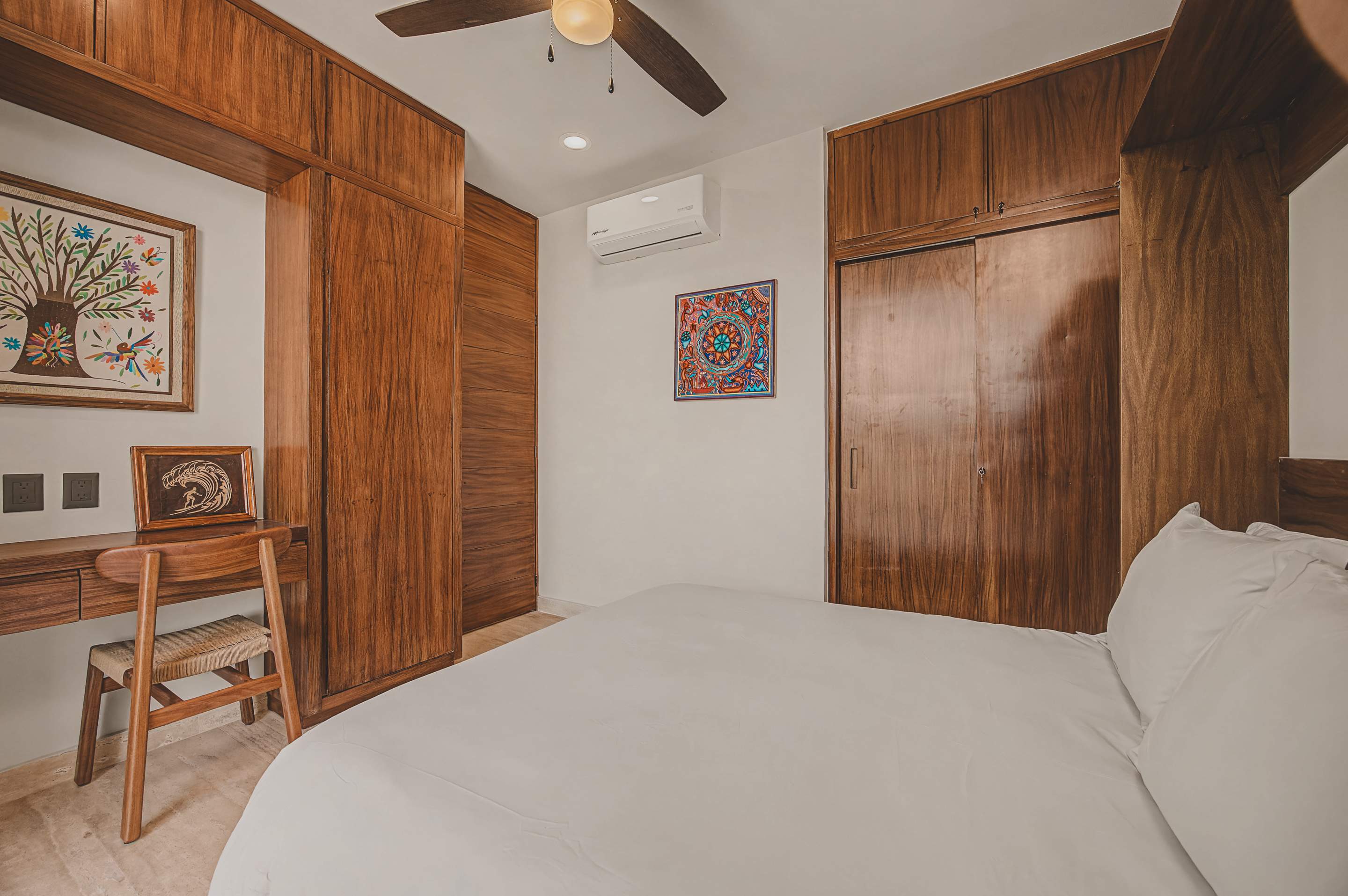 Modern bedroom with a white bed, extensive built-in wooden cabinetry and shelving, a small desk, and colorful artwork, with an air conditioning unit and ceiling fan visible.