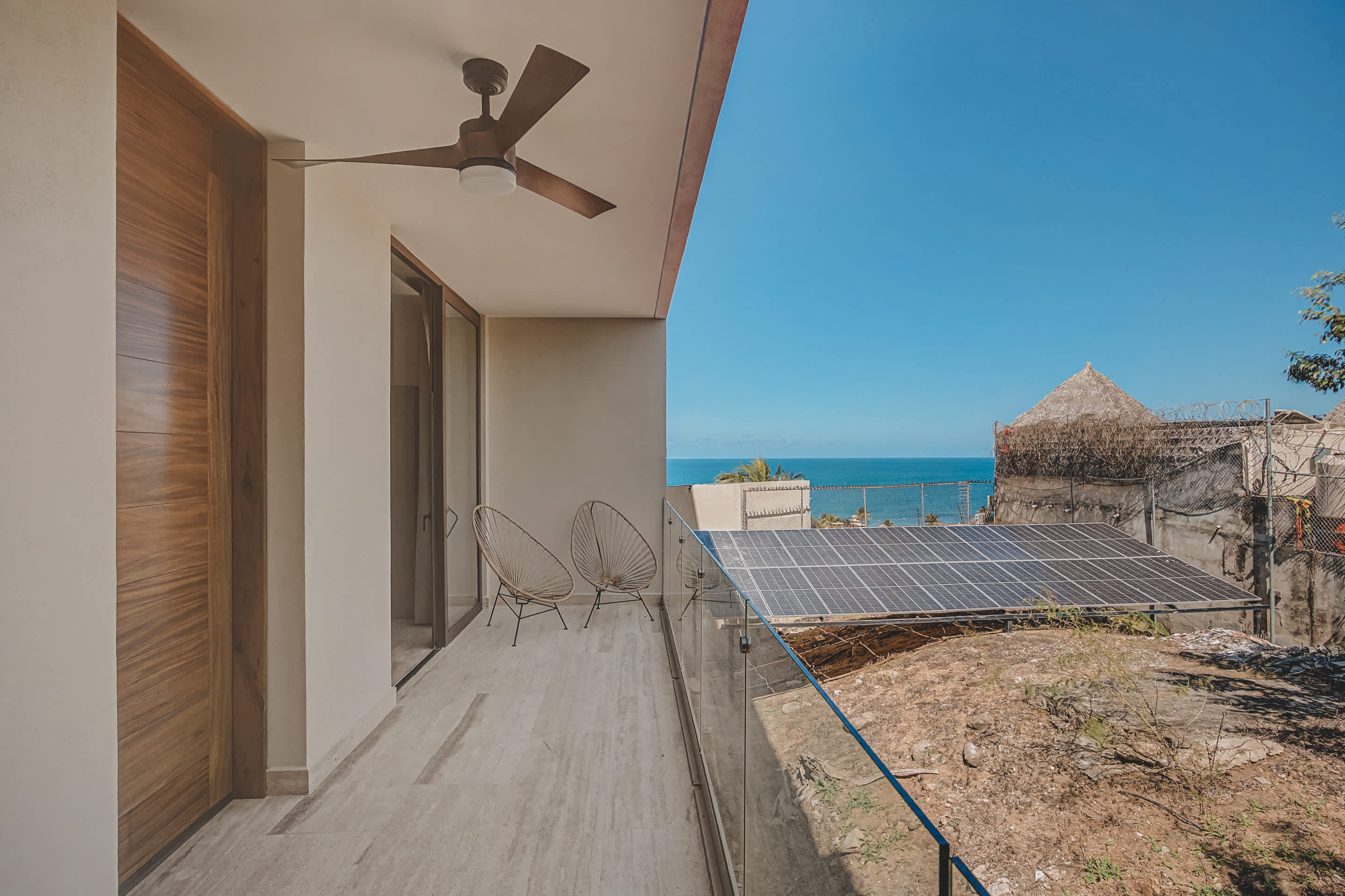 Spacious balcony with light-colored tiled flooring, a glass railing, and a ceiling fan, providing an expansive view of the ocean, solar panels, and rustic buildings under a bright blue sky.