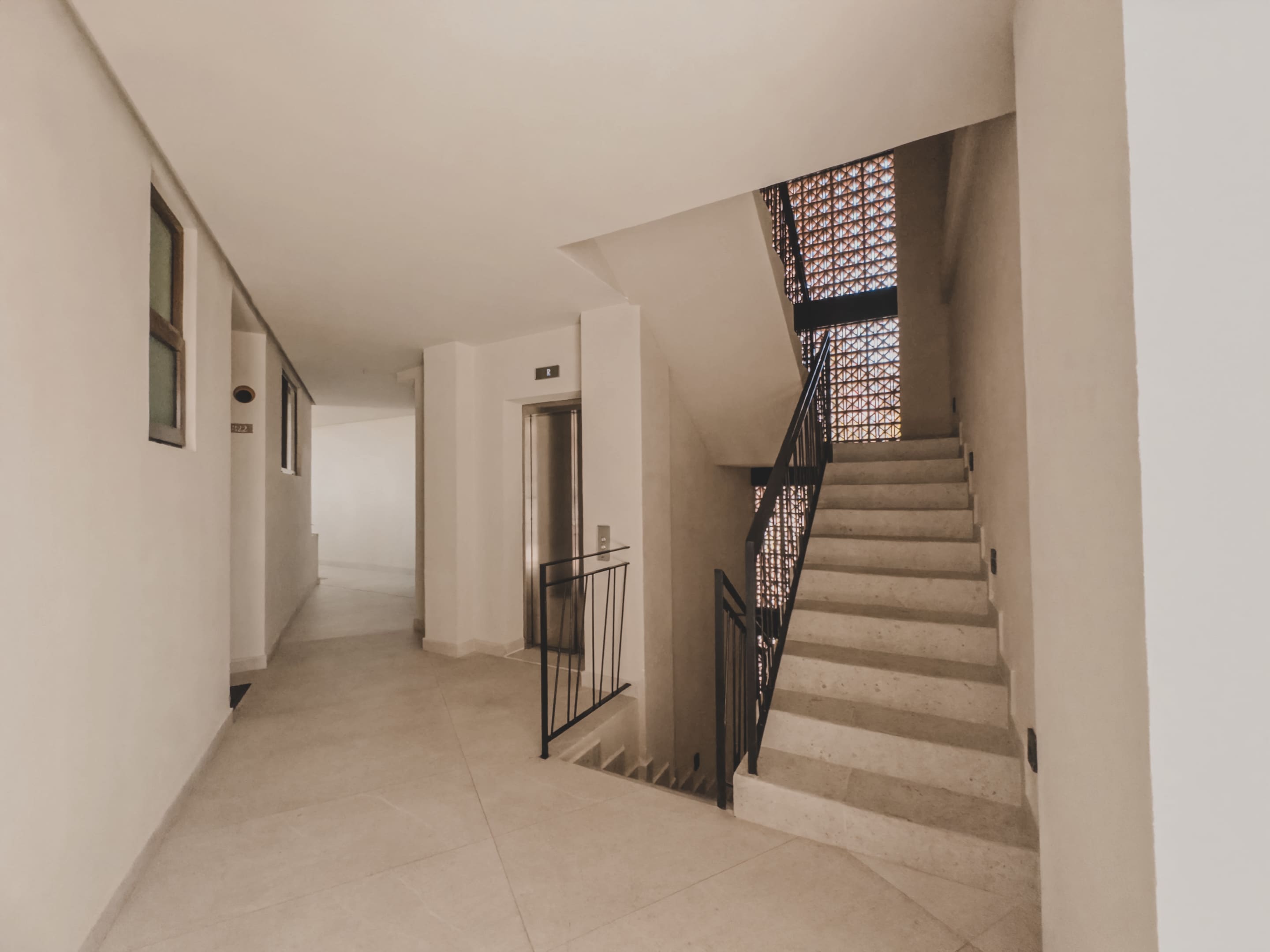 Bright interior hallway with light-colored tiled flooring, featuring a modern staircase with a black metal railing and an elevator, leading to various units in a contemporary building.