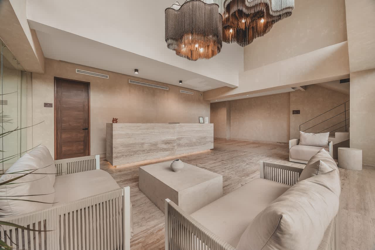 Inviting foyer with a long, light-colored stone reception desk and a comfortable lounge area featuring modern sofas and armchairs. Two large, decorative chandeliers illuminate the high-ceilinged space, which has light-toned walls and floors.