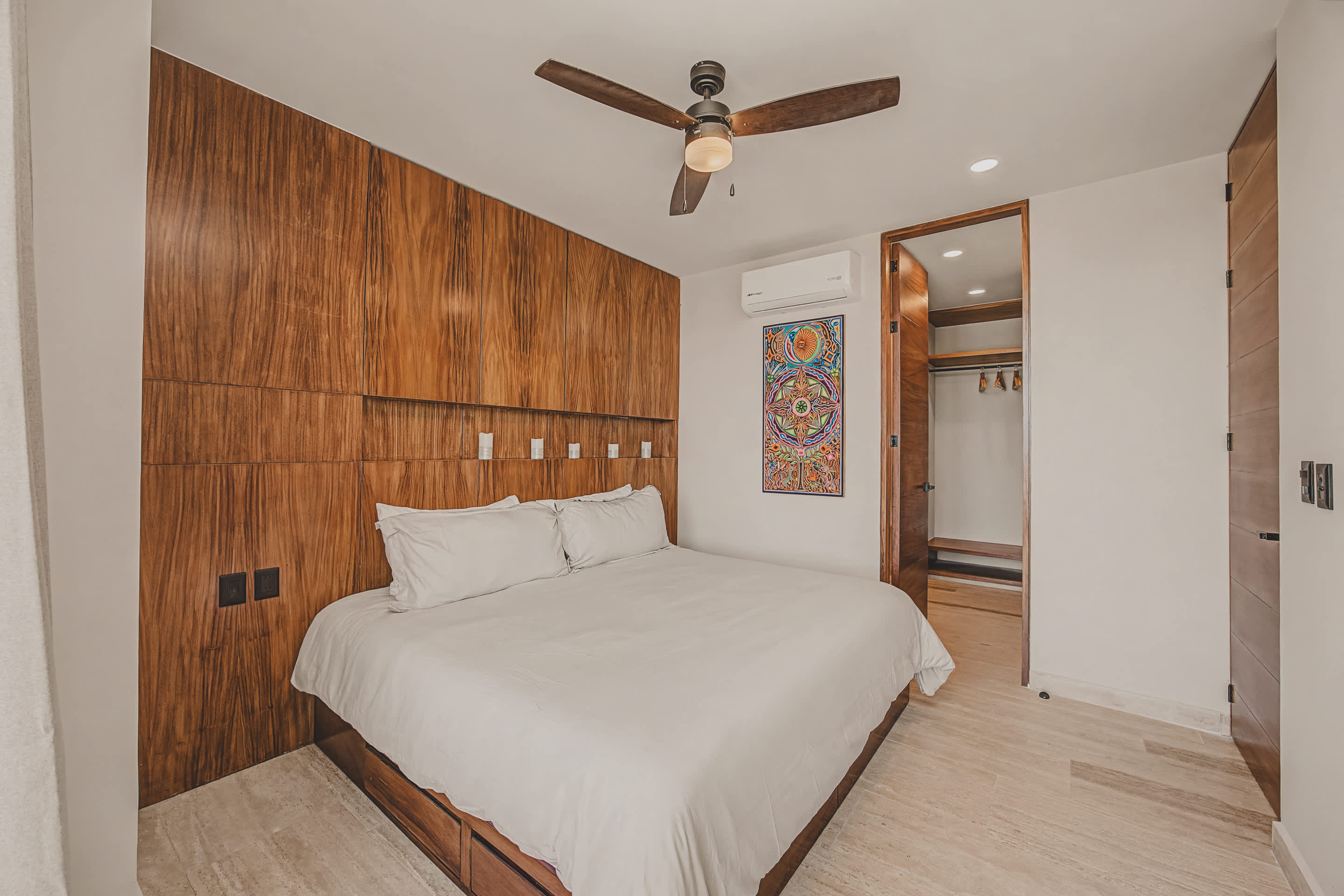 Spacious bedroom featuring a large white bed with a dark wooden platform and a full-height wooden panel headboard, complemented by a ceiling fan and colorful artwork.