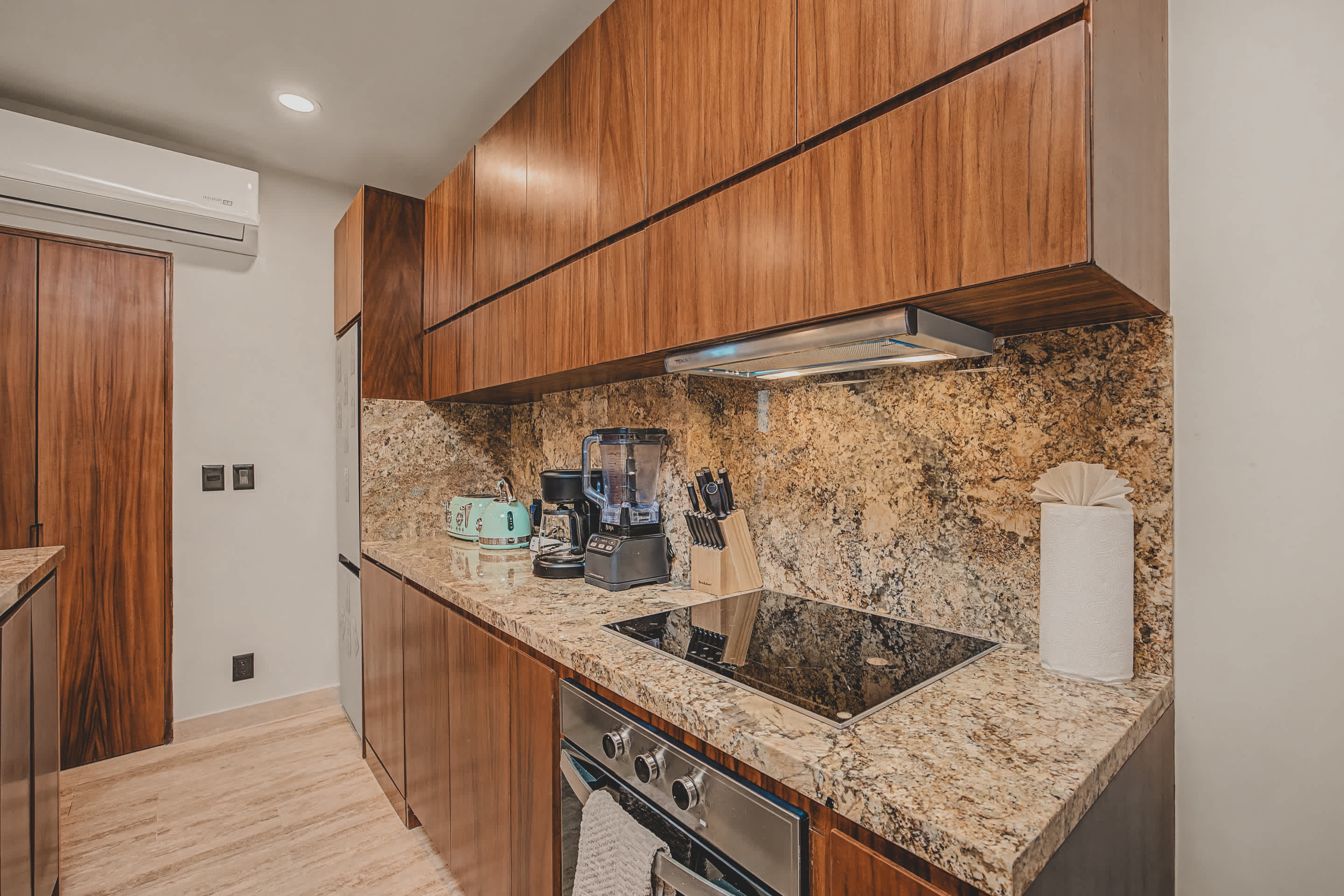 A well-equipped kitchen counter features speckled granite countertops and rich wood cabinetry, housing a modern induction cooktop and oven. Appliances like a coffee maker, blender, and a retro-style toaster and kettle are neatly arranged, with a roll of paper towels nearby.