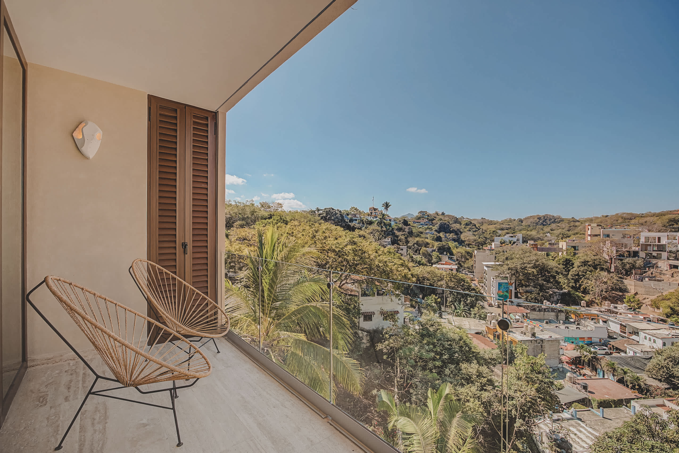 A balcony with two woven chairs offers a scenic view of a vibrant town and lush green hills under a bright blue sky. The modern building facade with wooden shutters is visible on the left.