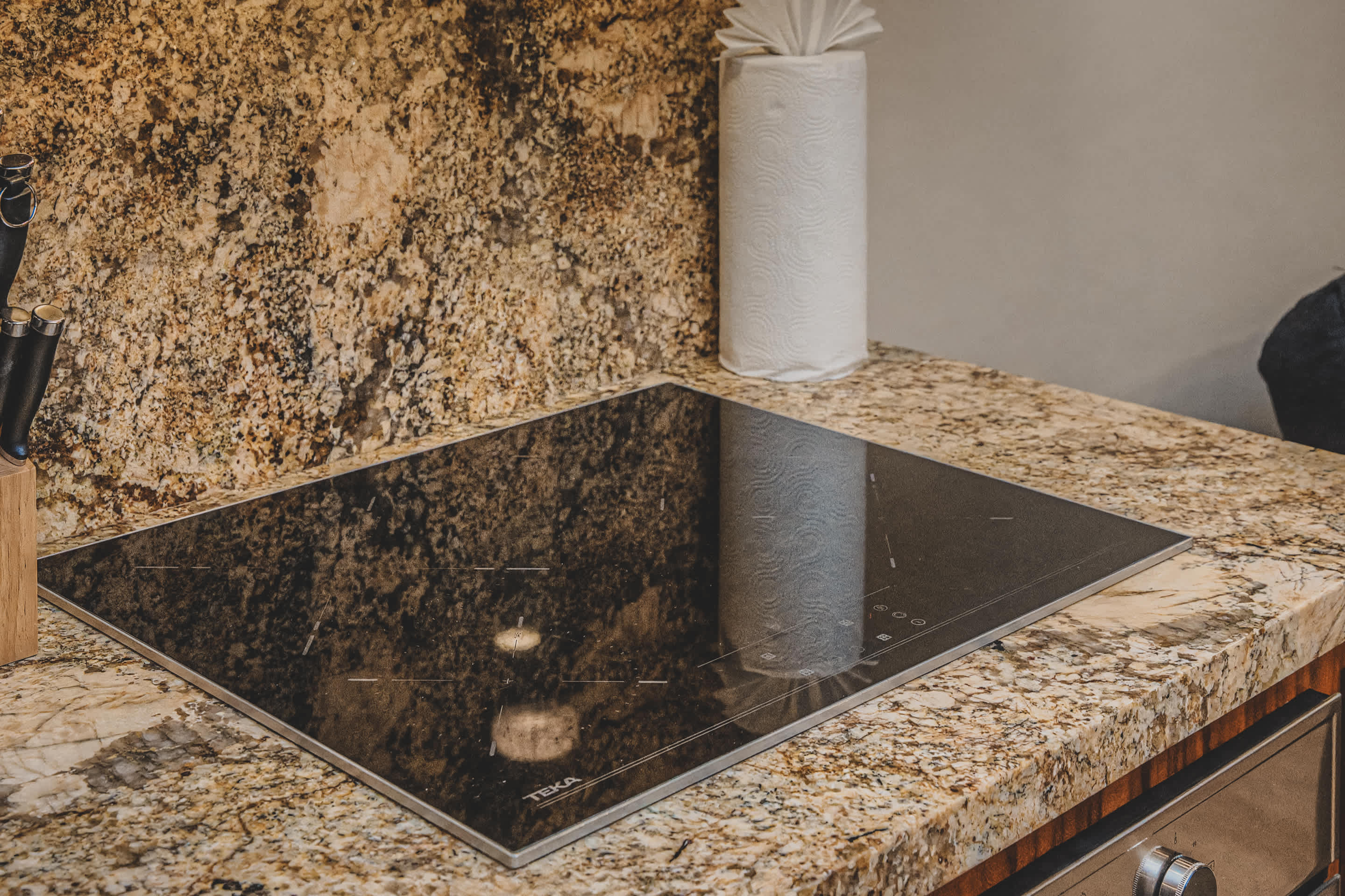Close-up of a modern black induction cooktop set into a speckled granite countertop, with a roll of paper towels and a knife block visible in the background.