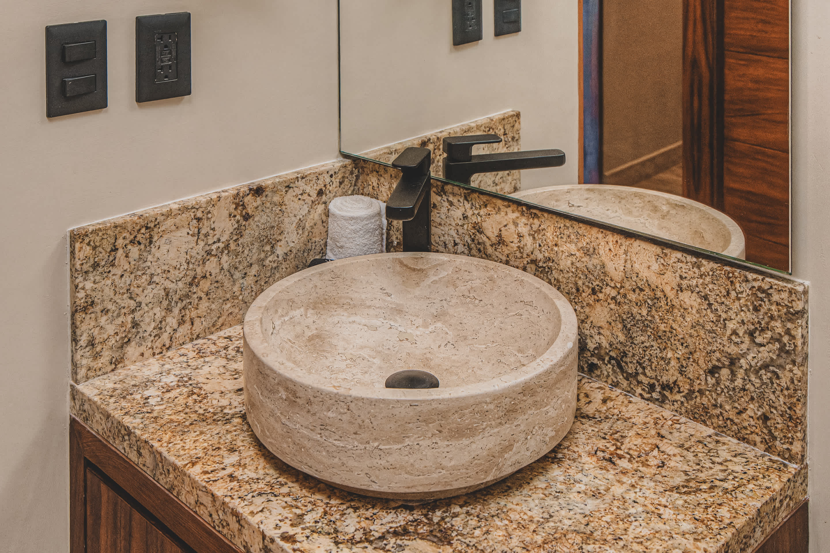 Bathroom vanity with a round stone vessel sink and a sleek black faucet, set on a speckled granite countertop with a large mirror reflecting the room and wall-mounted light switches.