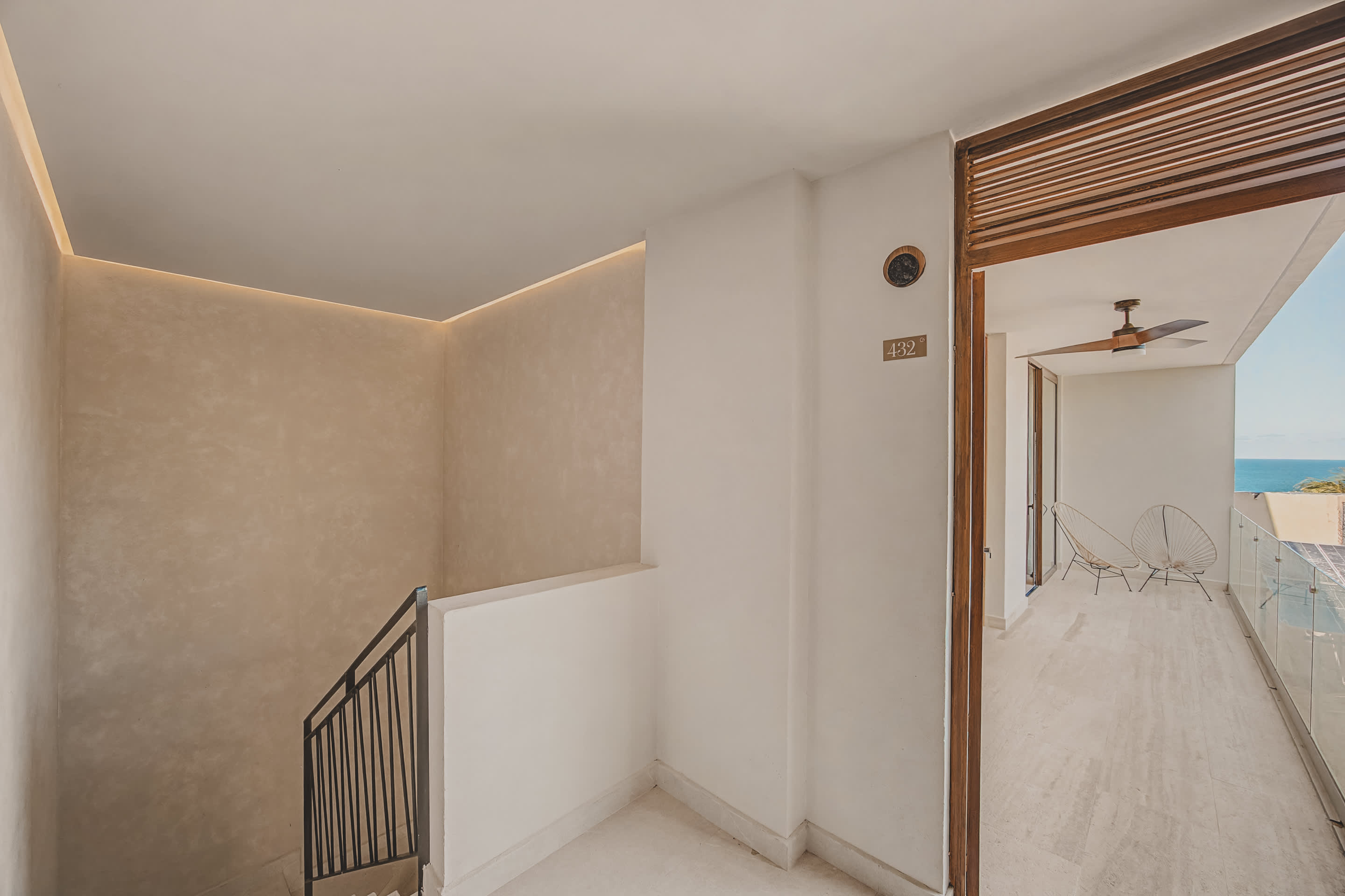 Minimalist interior foyer with light-colored walls and tiled flooring, featuring a modern staircase with a black metal railing and a wooden door leading to a balcony with an ocean view.
