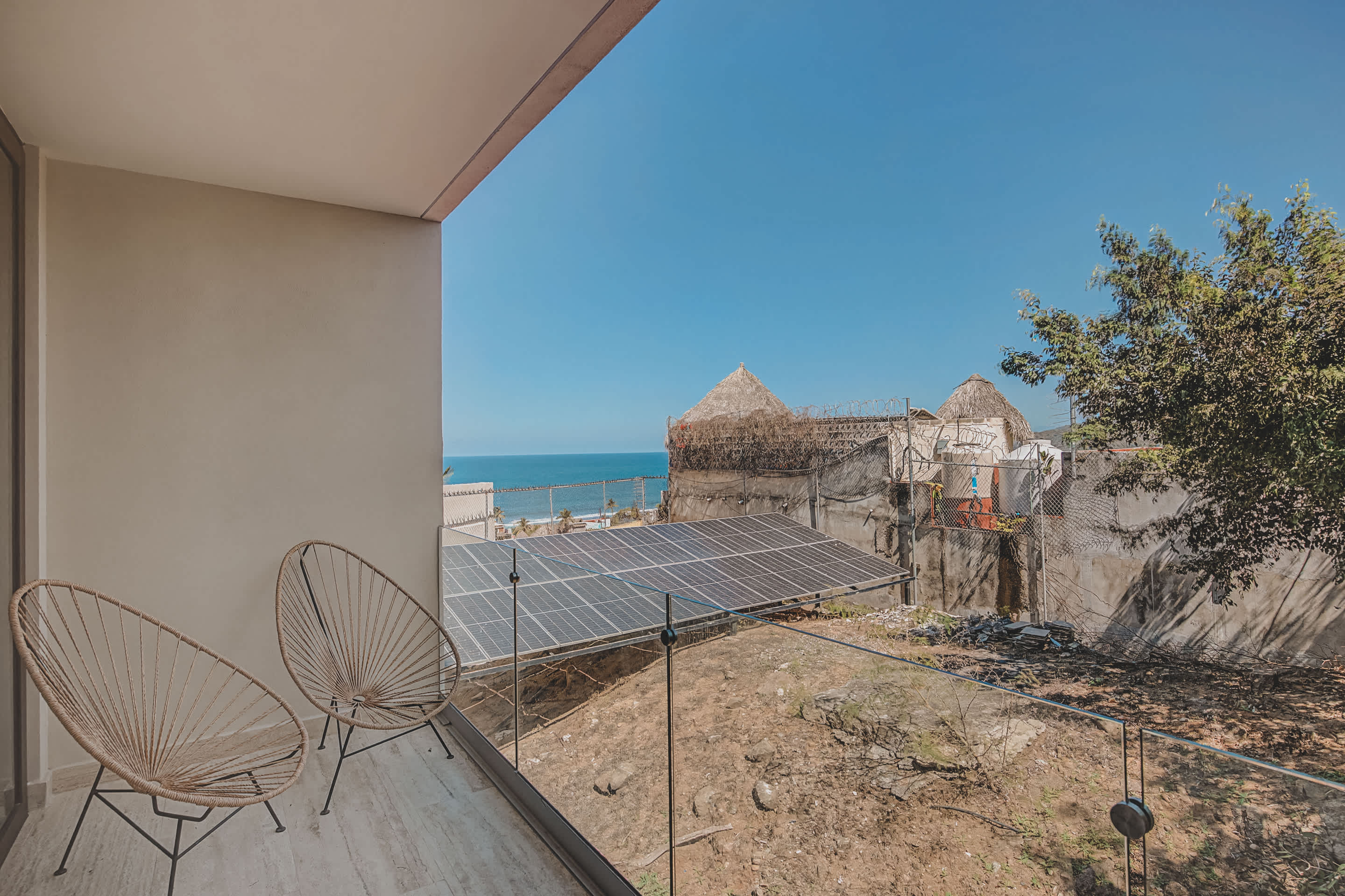A balcony with two woven chairs offers a partial view of the ocean and a nearby beach, with solar panels visible on a rooftop below. The foreground shows a somewhat undeveloped area with sparse vegetation.