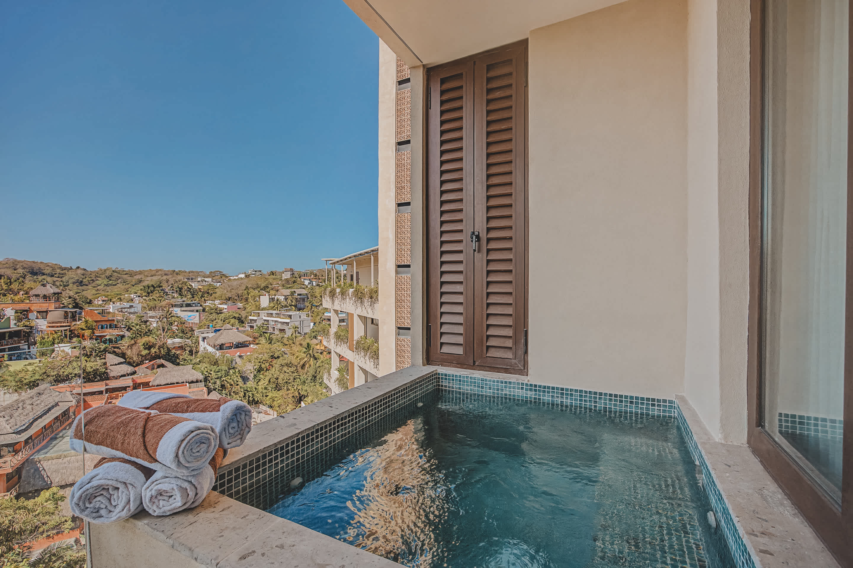 A close-up view of the private plunge pool on the balcony, featuring blue mosaic tiles and a stack of neatly folded towels. The background shows a modern building exterior and a clear blue sky.