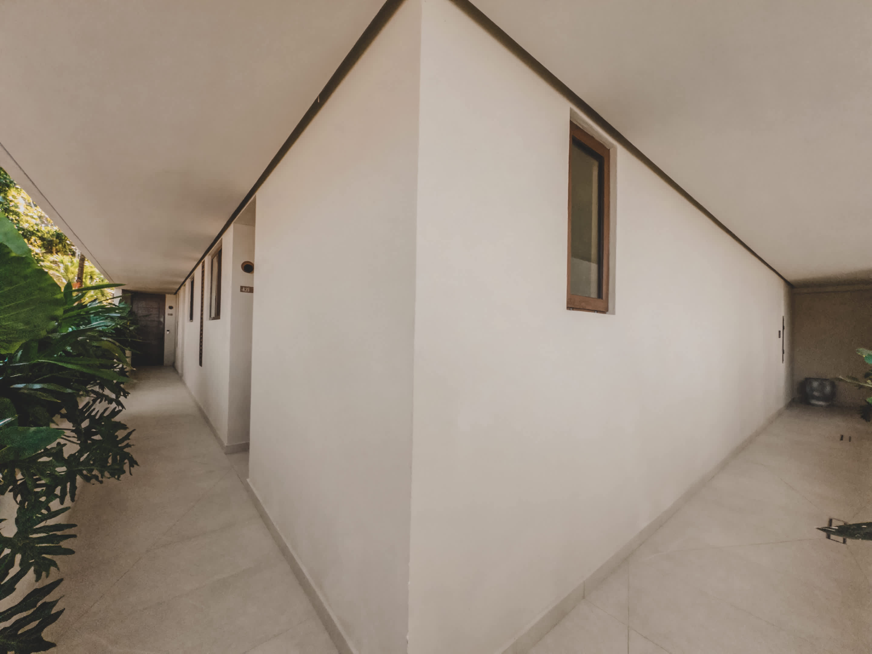 Outdoor hallway with light-colored tiled flooring and white walls, lined with lush green tropical plants, leading to multiple unit entrances under a covered walkway.