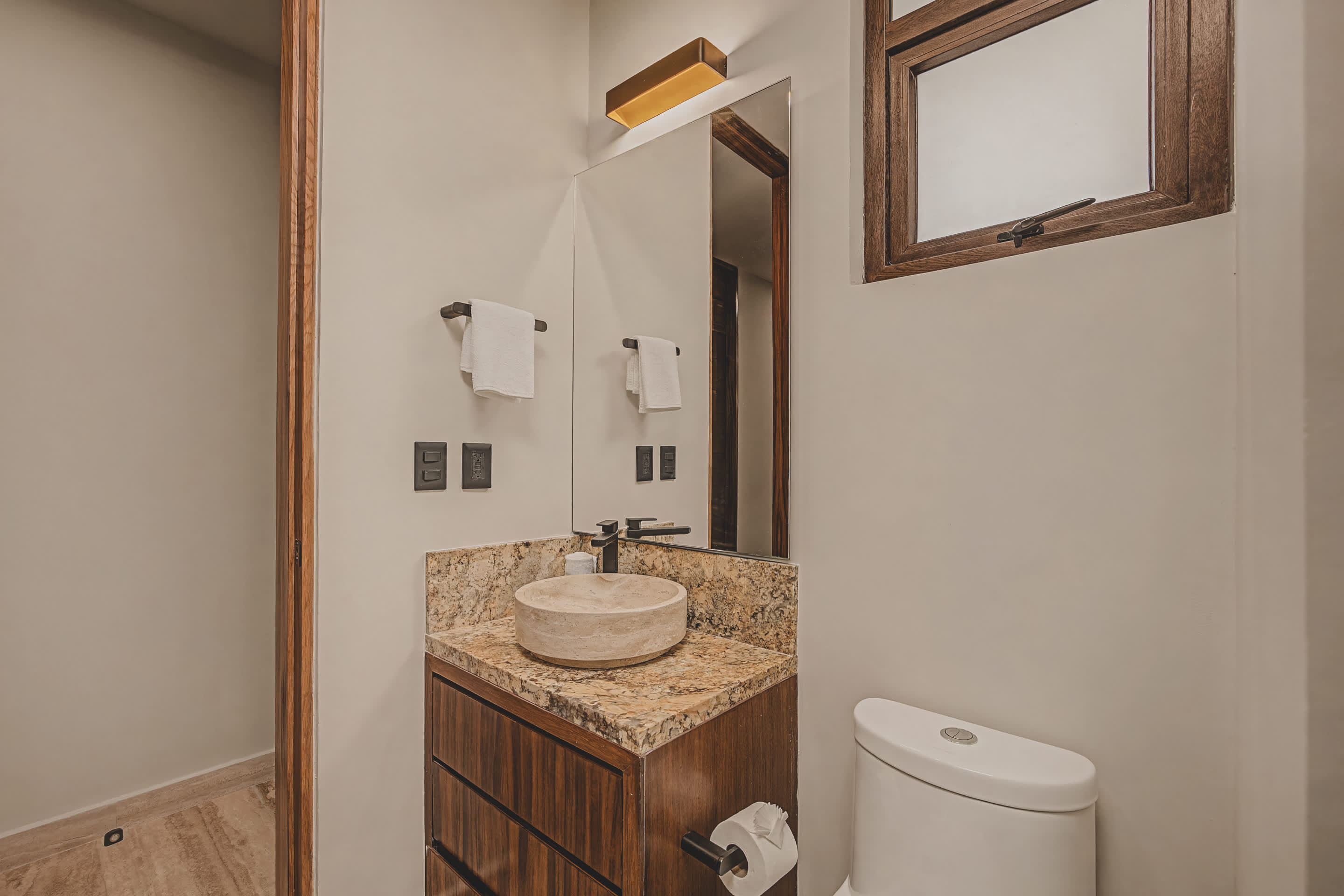 Modern bathroom with a light-colored vessel sink set on a speckled granite countertop, a dark wood vanity, a large mirror, and a toilet, complemented by a wooden window frame and dark fixtures.
