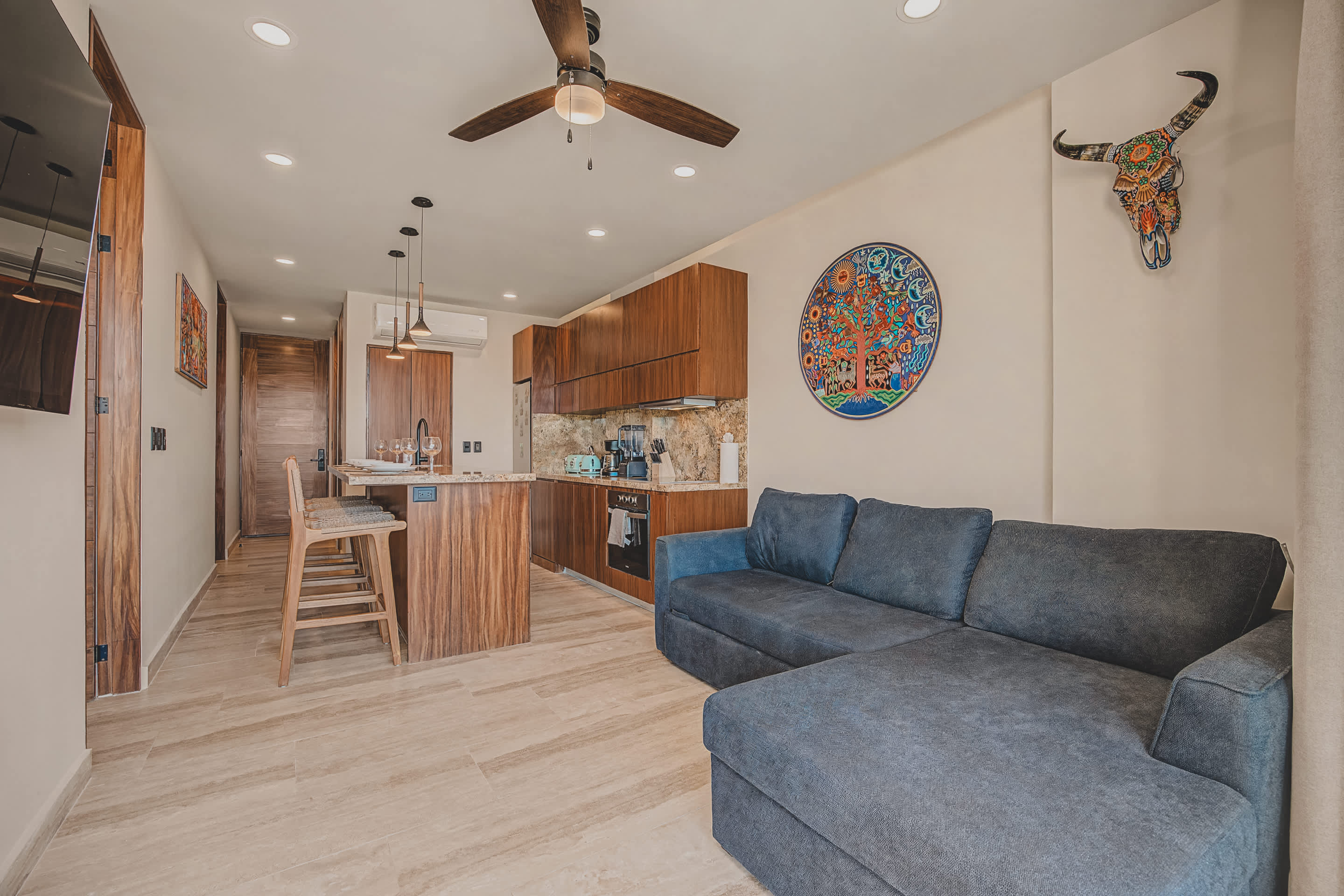 An inviting open-concept space combines a modern kitchen with dark wood cabinetry and a granite island, flowing into a living area with a dark sectional sofa. A colorful circular artwork adorns the wall, and a ceiling fan hangs above, with a glimpse of the balcony beyond.