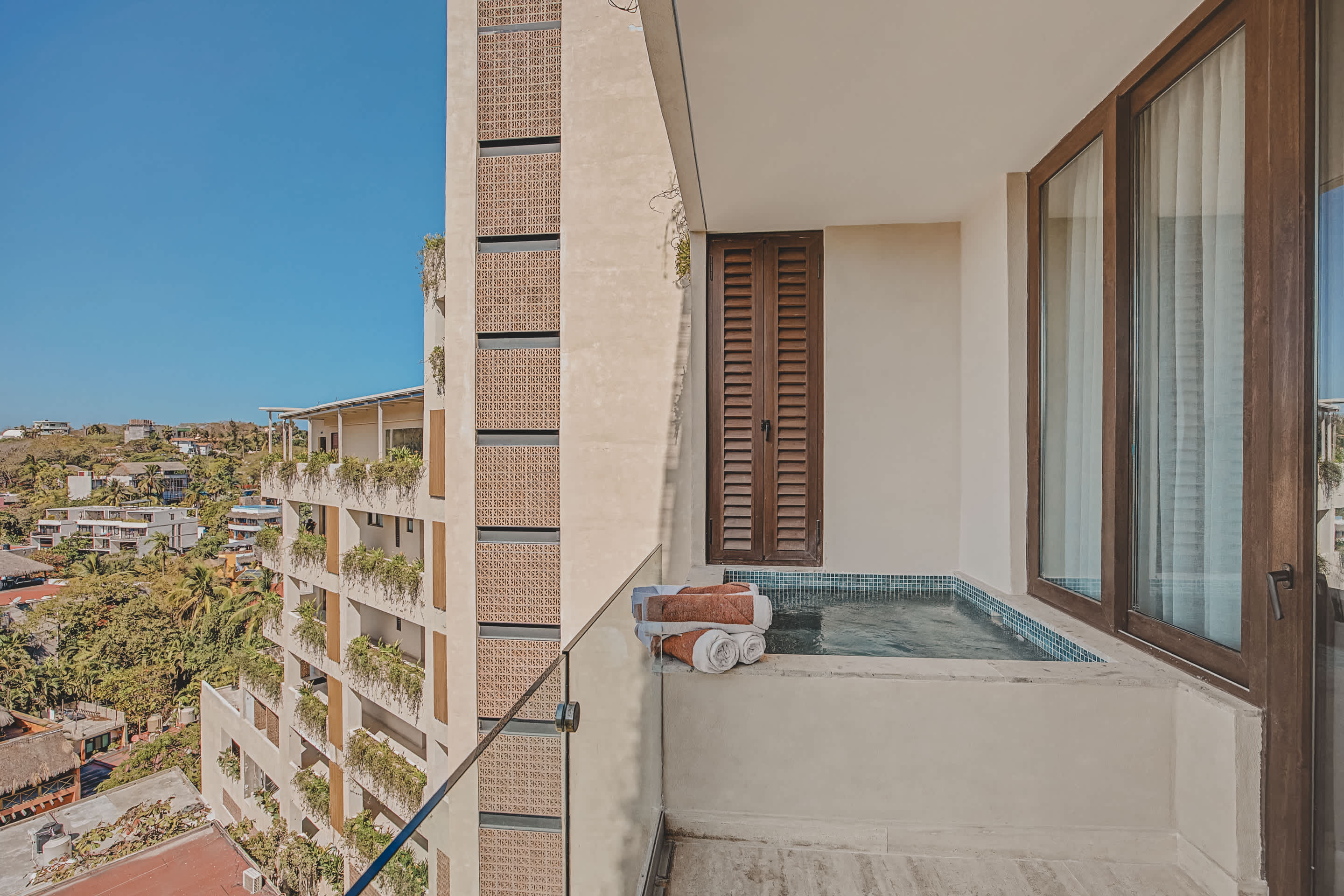This balcony features a private plunge pool with blue mosaic tiles and a stack of towels, set against a modern building facade. It offers a wide view of a bustling town nestled among lush green hills under a clear blue sky.