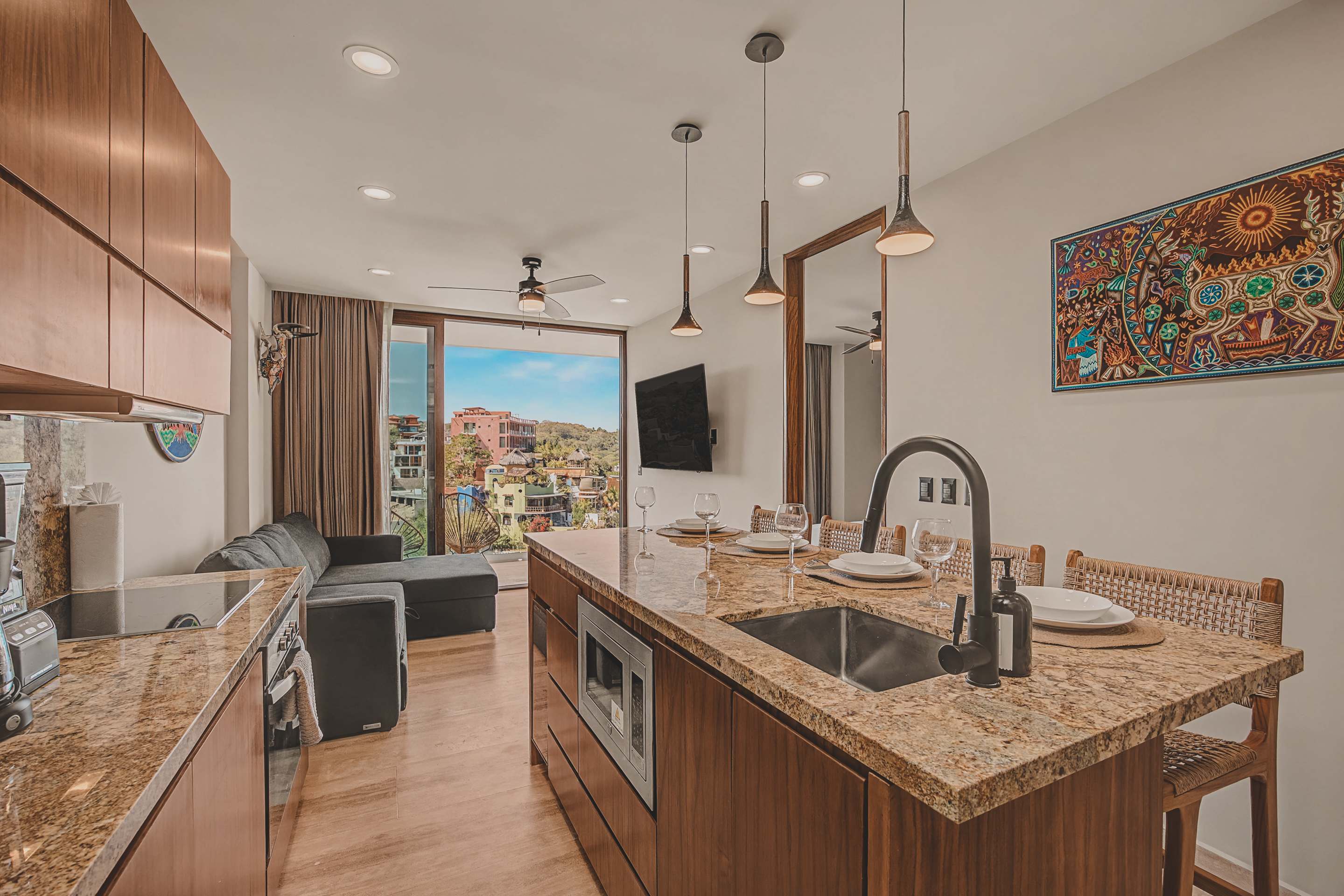 The sleek kitchen features dark wood cabinetry and speckled granite countertops, with a large island incorporating a sink and microwave, serving as a dining space. Modern pendant lights hang above the island, which is set for dining, with a view towards the living room and balcony.