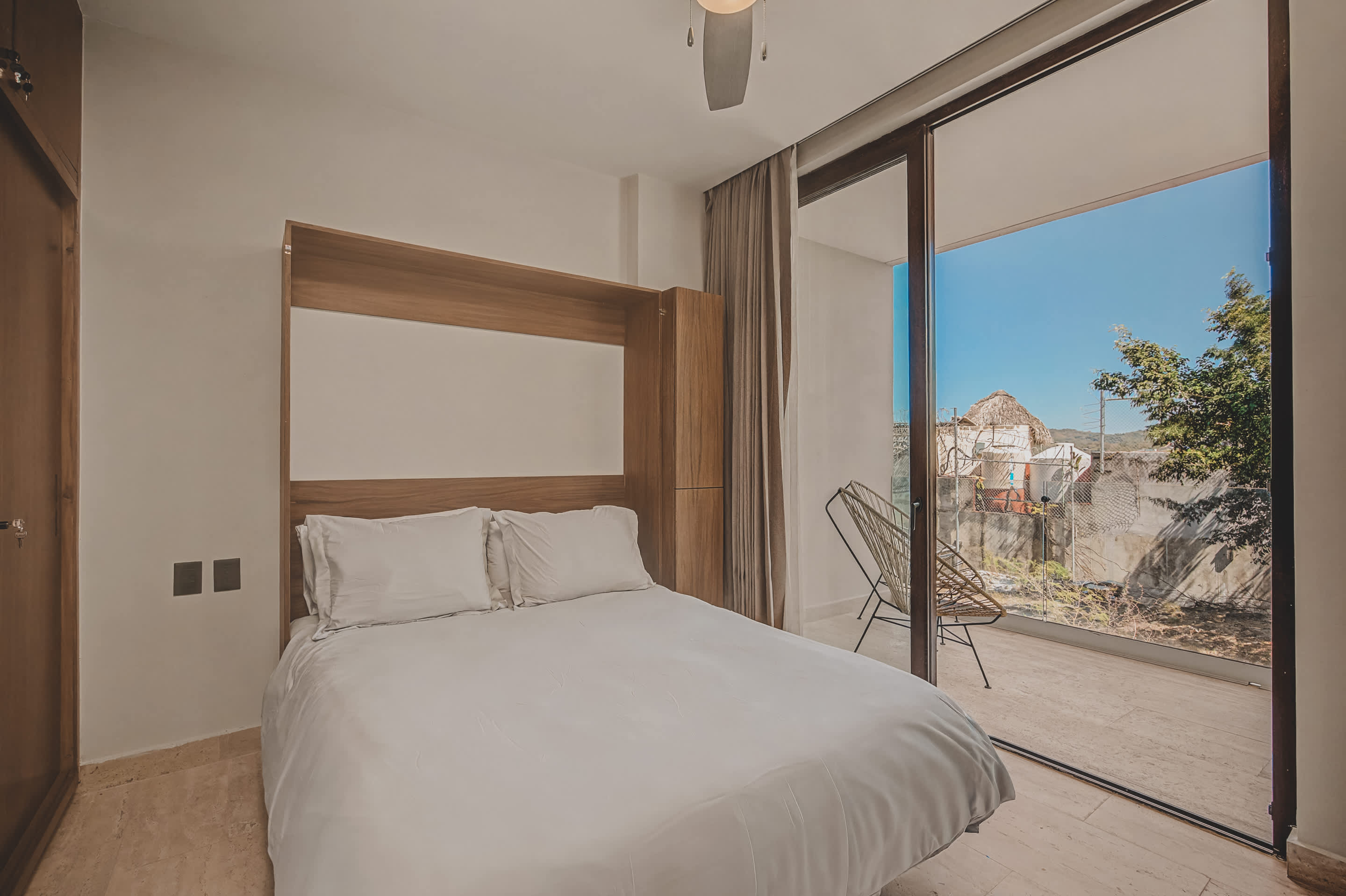 Bright bedroom with a white bed integrated into a light wooden wall unit, featuring large sliding glass doors that open to a private balcony with outdoor seating and a view of trees.