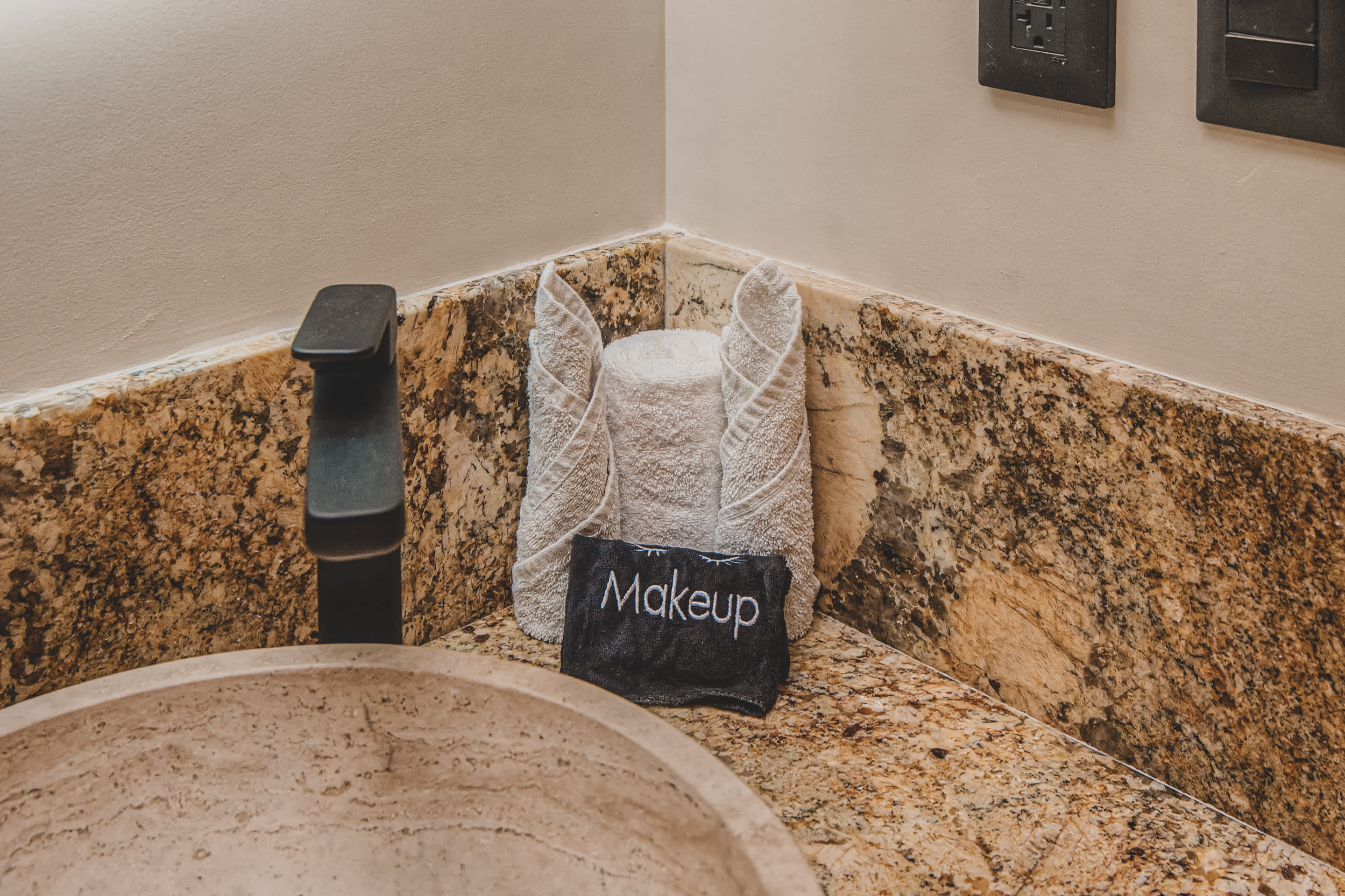 Close-up of a speckled granite bathroom countertop featuring a black faucet, a stack of neatly folded white towels, and a small black "Makeup" bag next to a stone vessel sink.