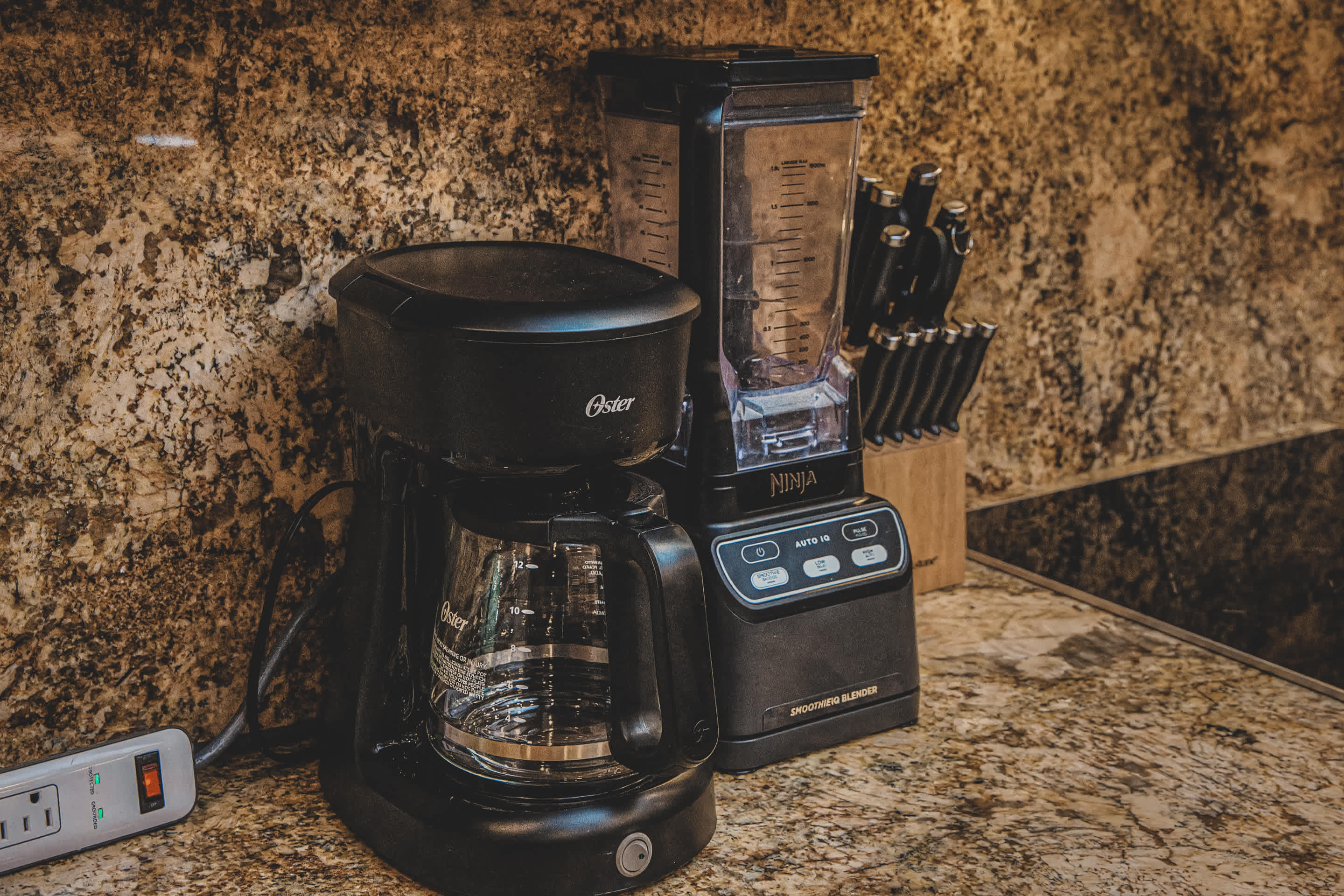 Kitchen countertop featuring a black Oster coffee maker and a Ninja smoothie blender, set against a speckled granite backsplash and countertop, with a knife block visible in the background.