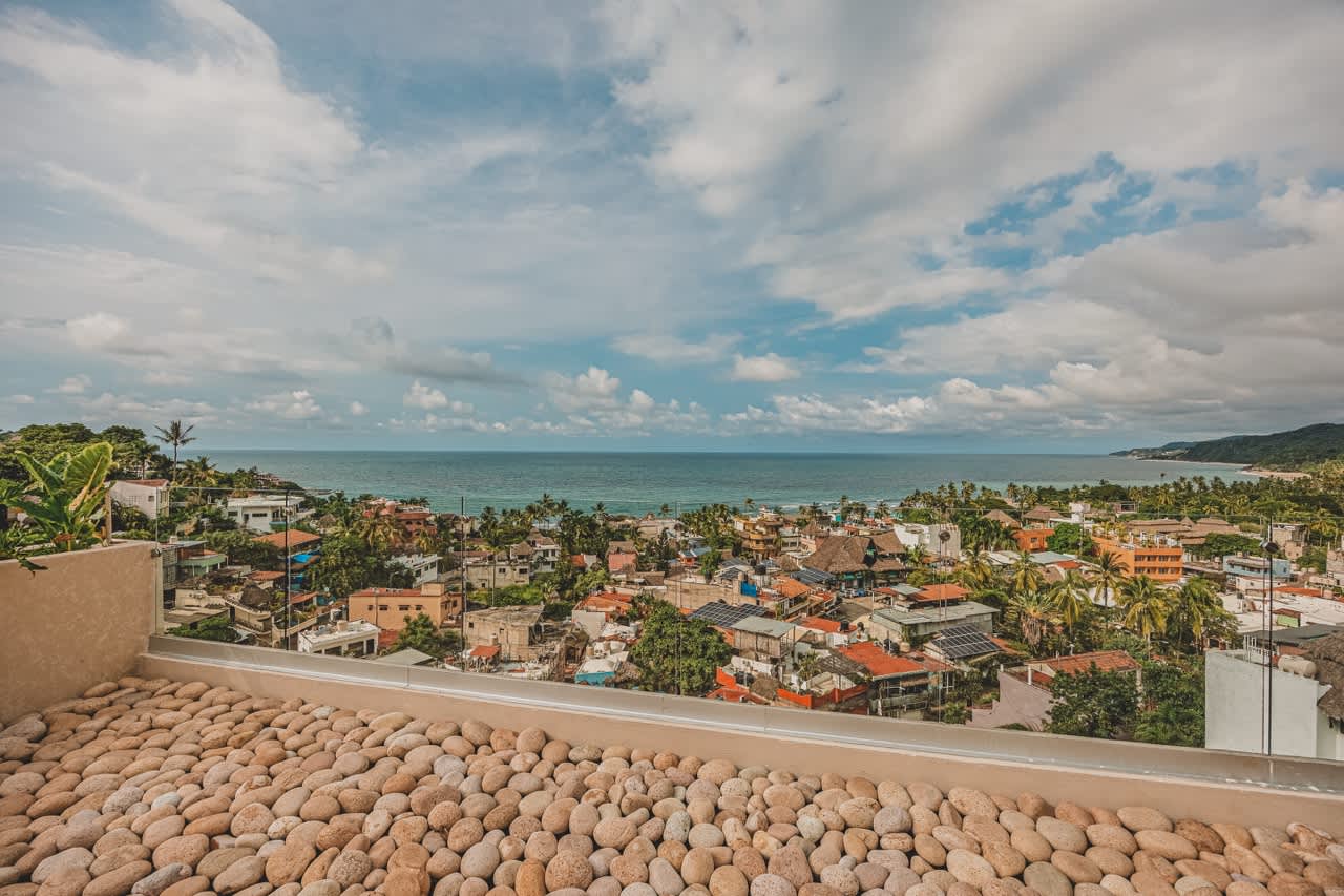 Expansive rooftop deck covered in light-colored pebbles, providing a sweeping panoramic view of the ocean, coastline, and a vibrant coastal town under a cloudy sky.