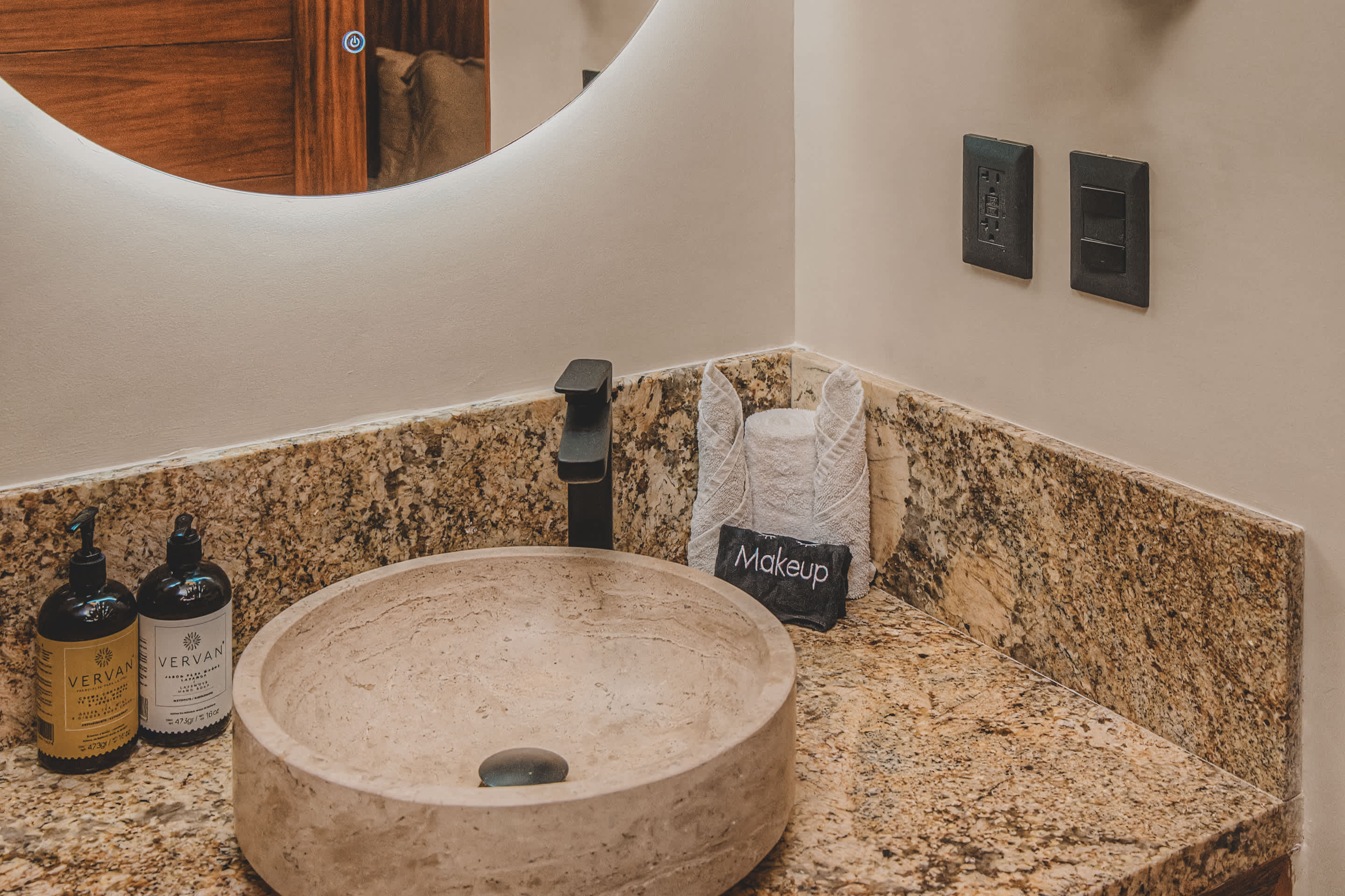 Modern bathroom vanity with a round, light-colored stone vessel sink, a black faucet, and speckled granite countertops, with two bottles of toiletries and a mirror reflecting the room.