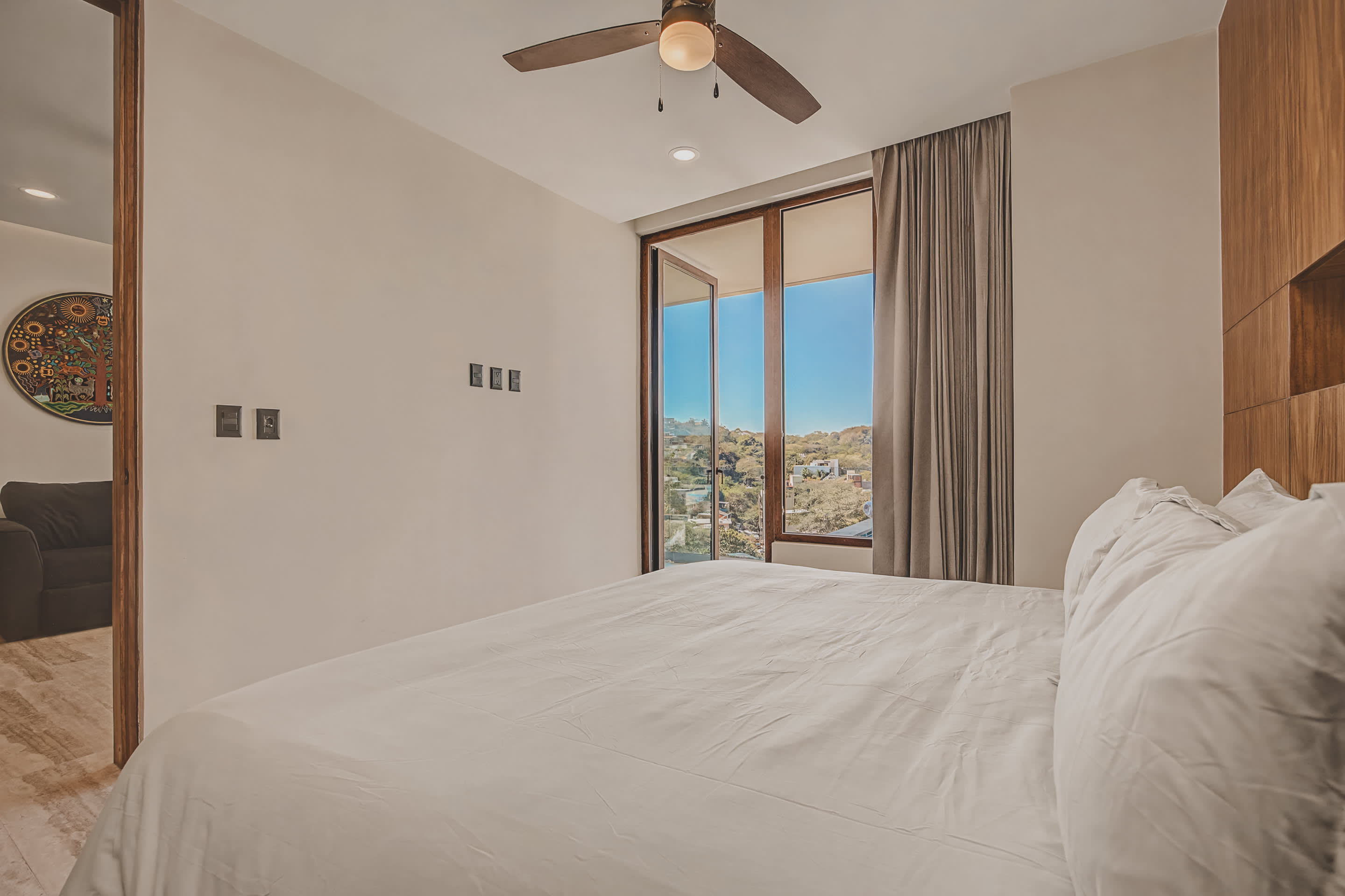 A bright bedroom features a large bed with white linens and a ceiling fan, with light-colored walls and a wooden accent panel. A large window with drapes offers a view of the vibrant cityscape and hills, bringing natural light into the room.
