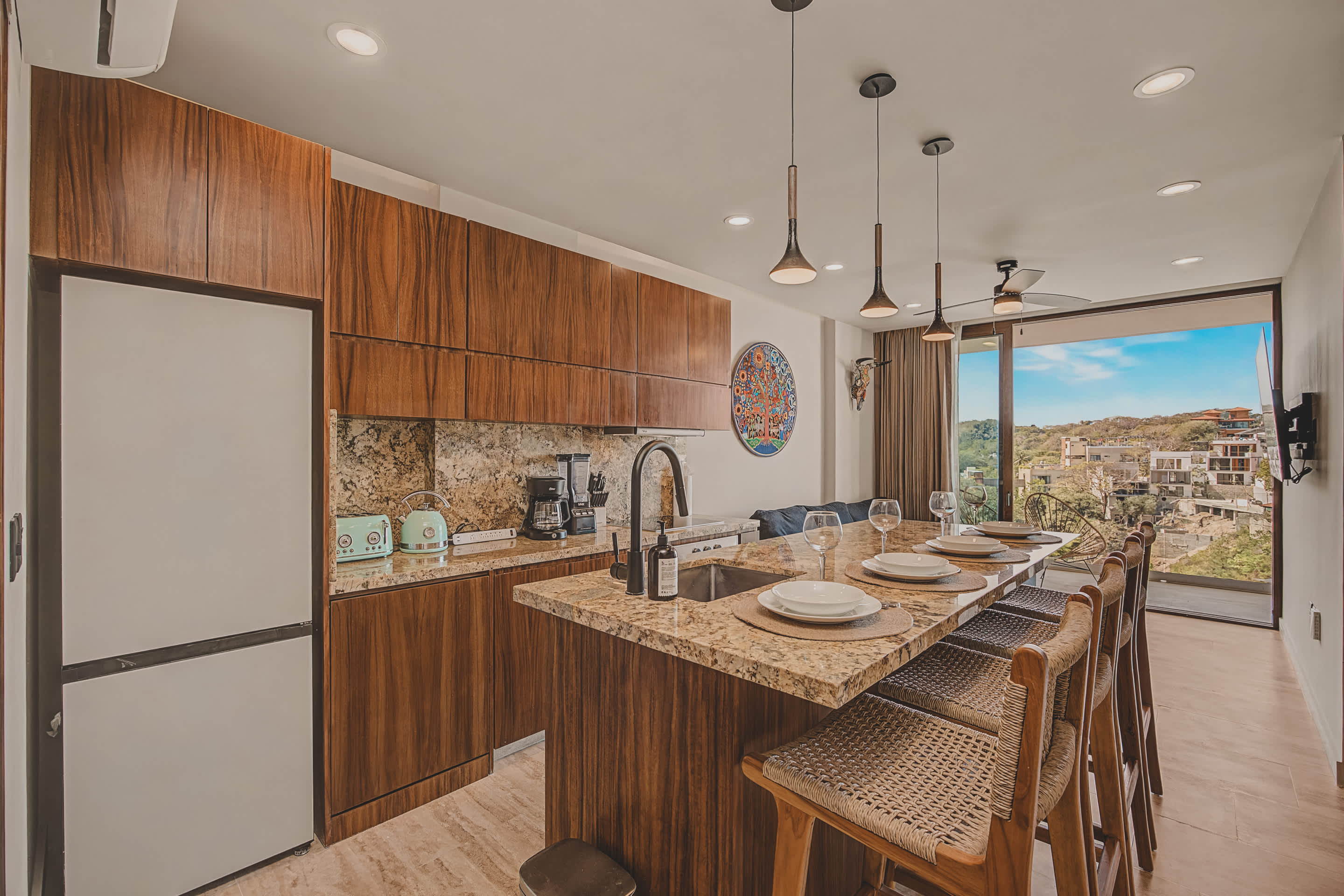 A modern kitchen boasts rich wood cabinetry, granite countertops, and stainless steel appliances, with a large island serving as a dining bar. Pendant lights illuminate the island, which is set with plates and glasses for four, overlooking the living area and balcony view.