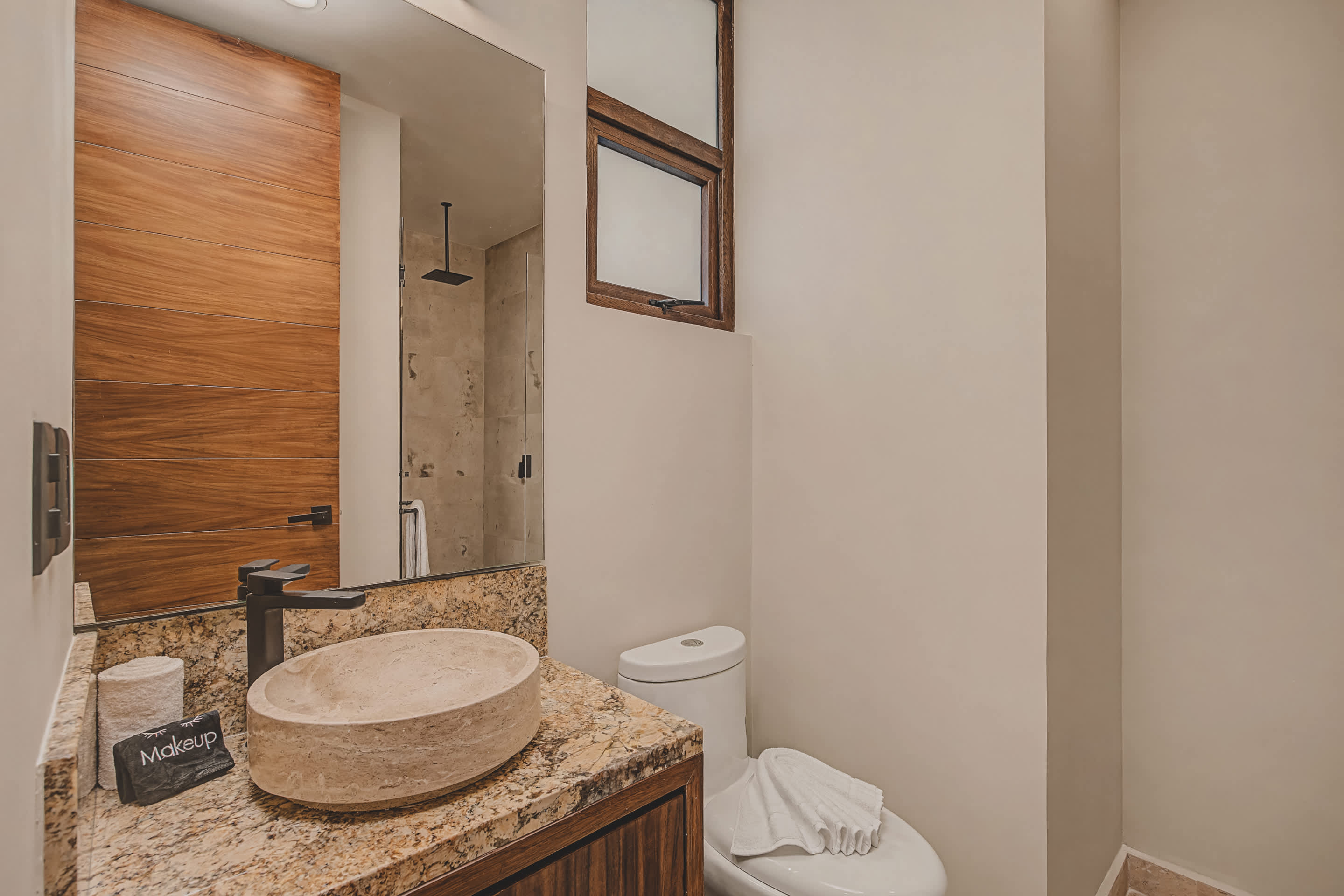 Contemporary bathroom featuring a light-colored vessel sink on a granite countertop with a dark wood vanity, a large mirror, and a toilet, with a wooden window frame above.