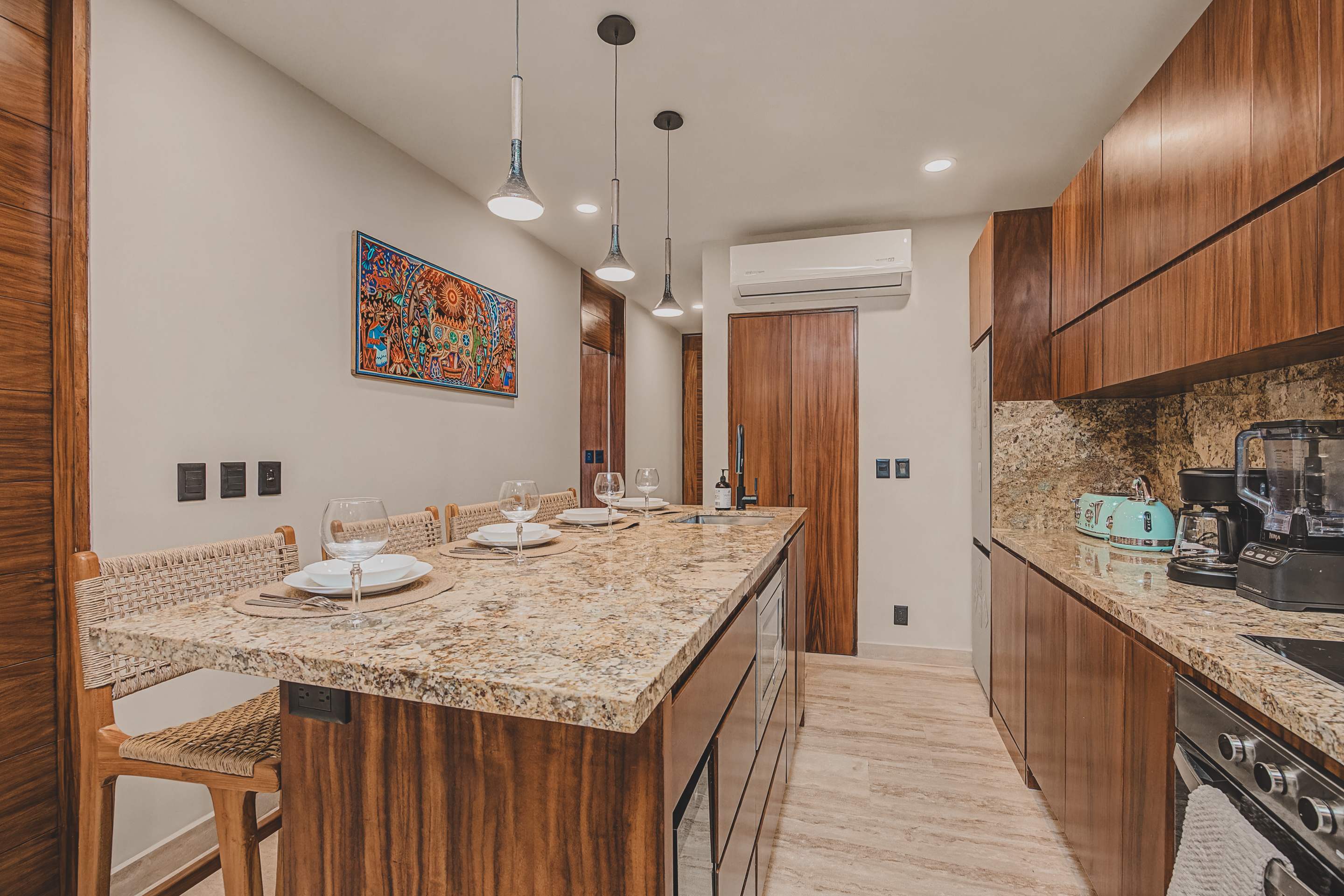 The contemporary kitchen features dark wood cabinetry and granite countertops, with a long island serving as a dining bar, set with four woven stools and place settings. Modern pendant lights illuminate the space, and a colorful artwork hangs on the wall, adding a vibrant touch.