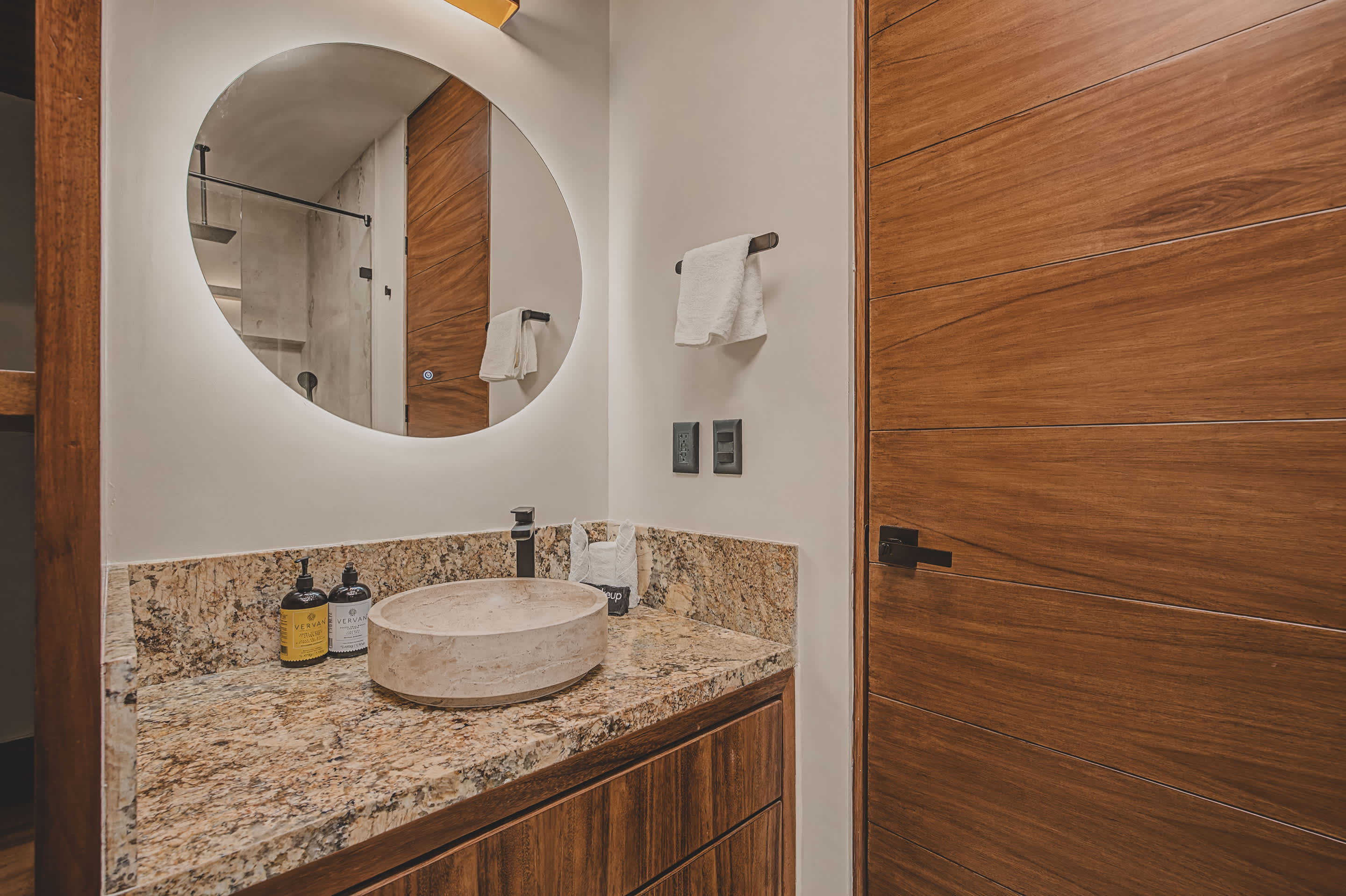 Detailed view of a stylish bathroom vanity featuring a light granite countertop, a unique stone vessel sink, and a large circular backlit mirror, with modern black fixtures and toiletries.