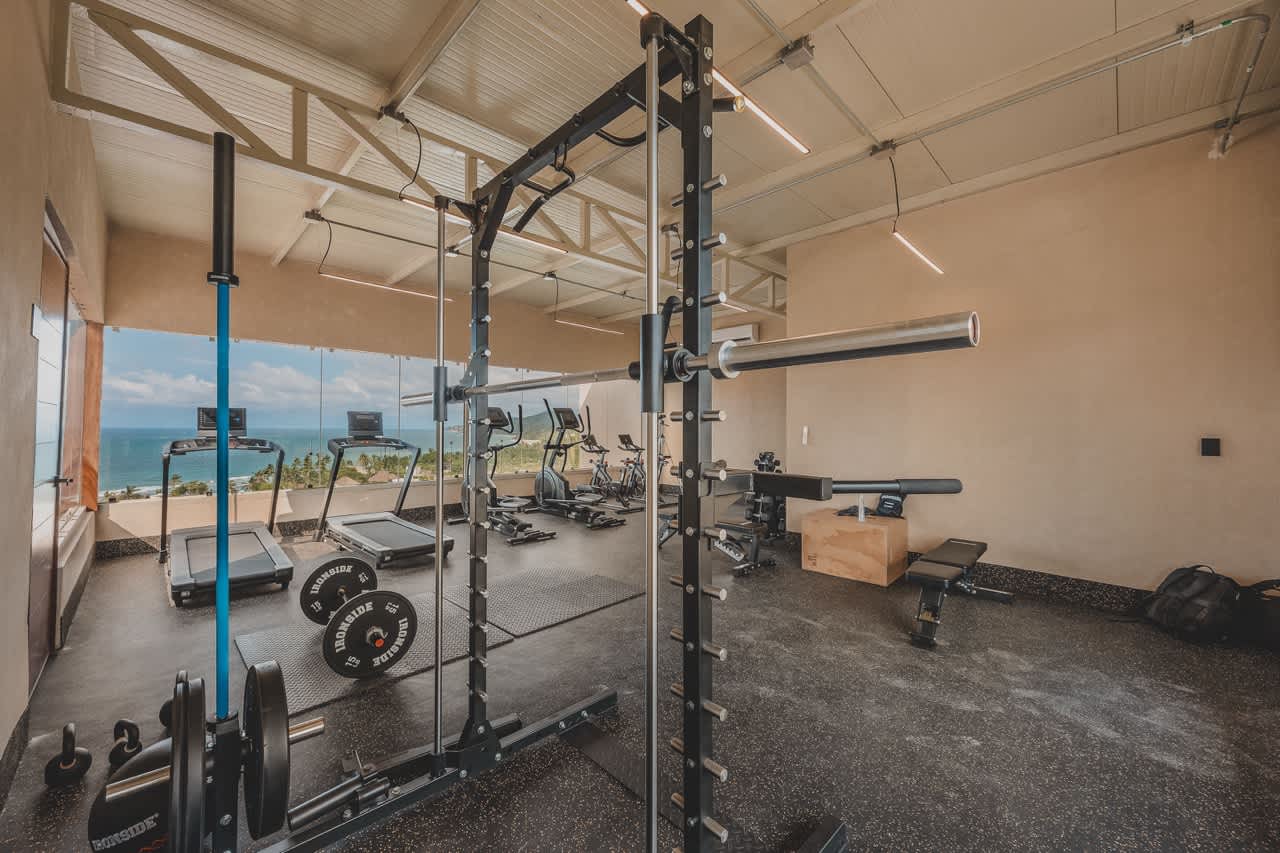 Spacious gym with a power rack, barbell, weight plates, and a bench, complemented by cardio machines in the background and a stunning ocean view through large windows. The ceiling has exposed white trusses.