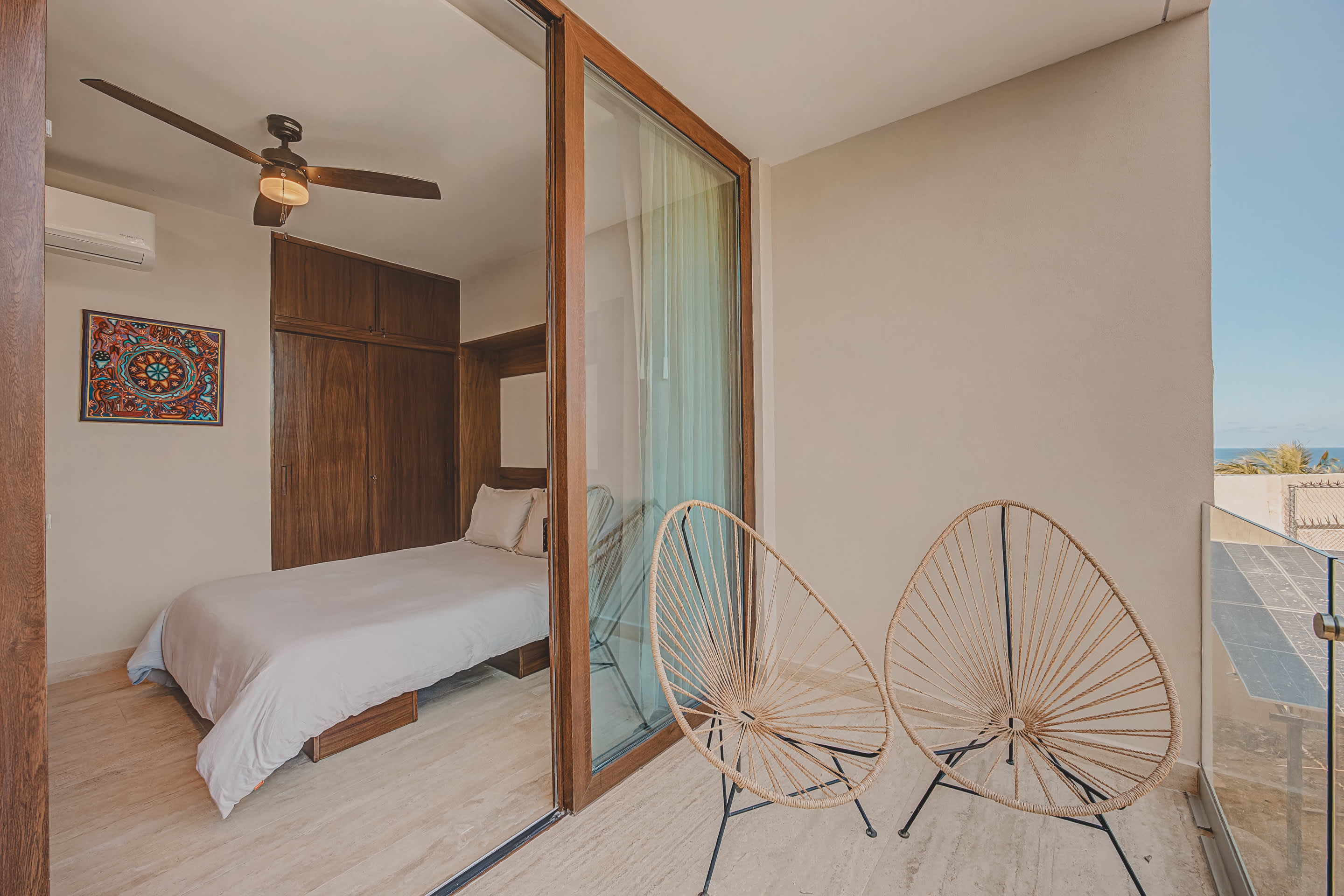 Bright bedroom with a white bed, dark wood built-in cabinetry, and a colorful framed artwork, visible through a large sliding glass door from the balcony with two woven chairs.