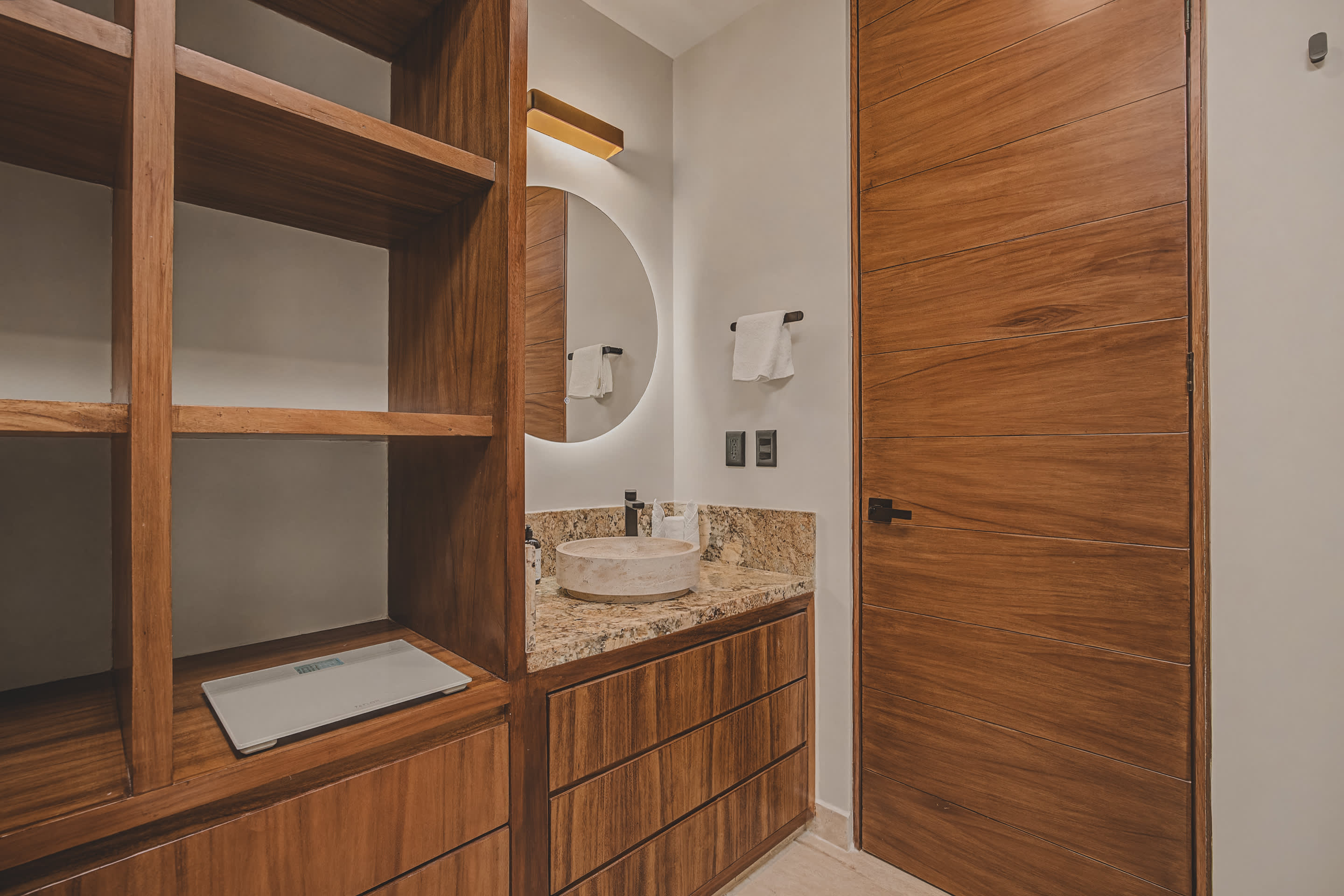 View of a modern bathroom vanity with a stone vessel sink and a large round backlit mirror, integrated into a spacious walk-in closet with extensive wooden shelving and a scale.