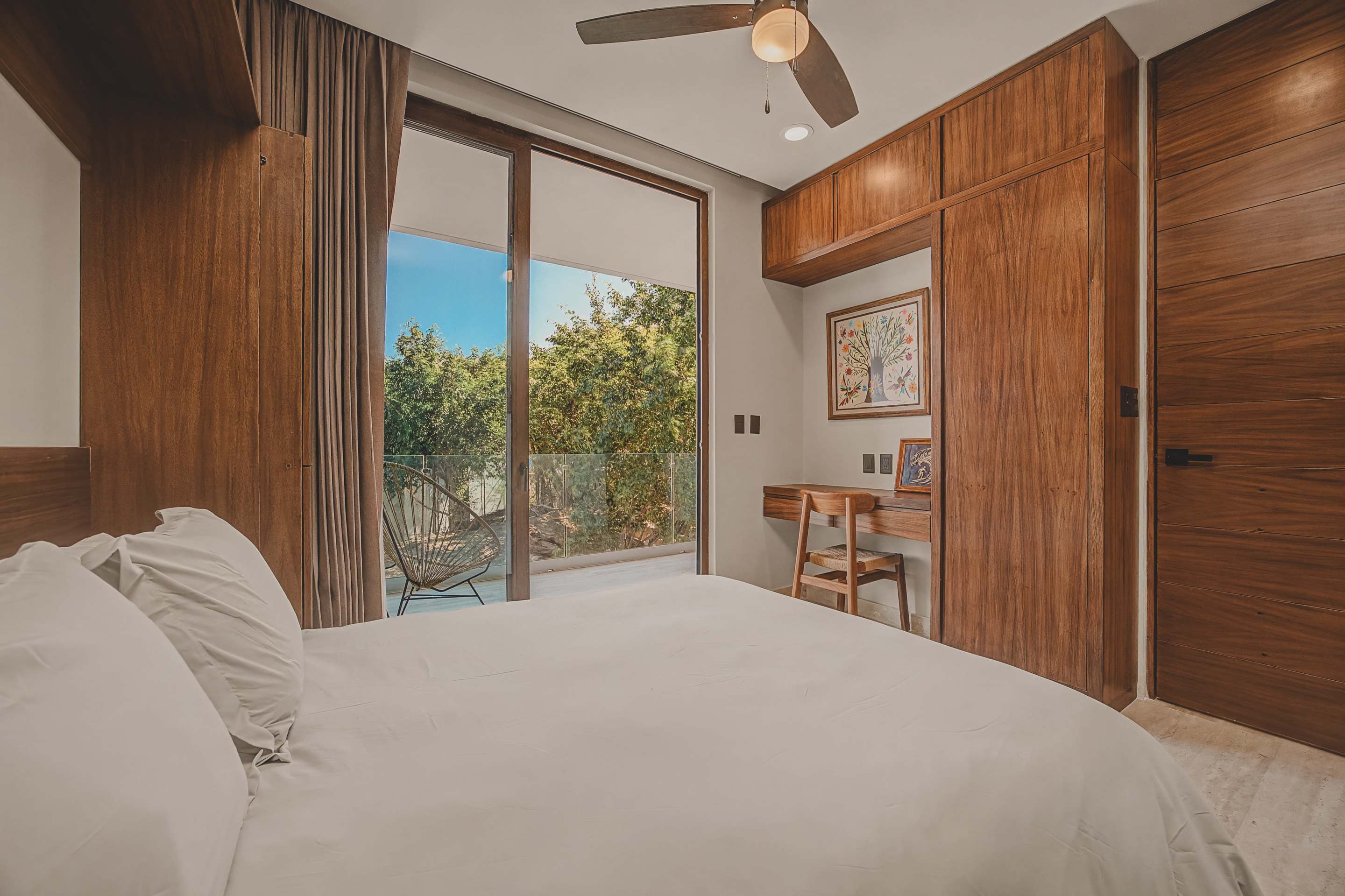 Cozy bedroom with a white bed, a ceiling fan, and large sliding glass doors leading to a balcony, featuring built-in wooden cabinetry, a small desk area, and colorful artwork.