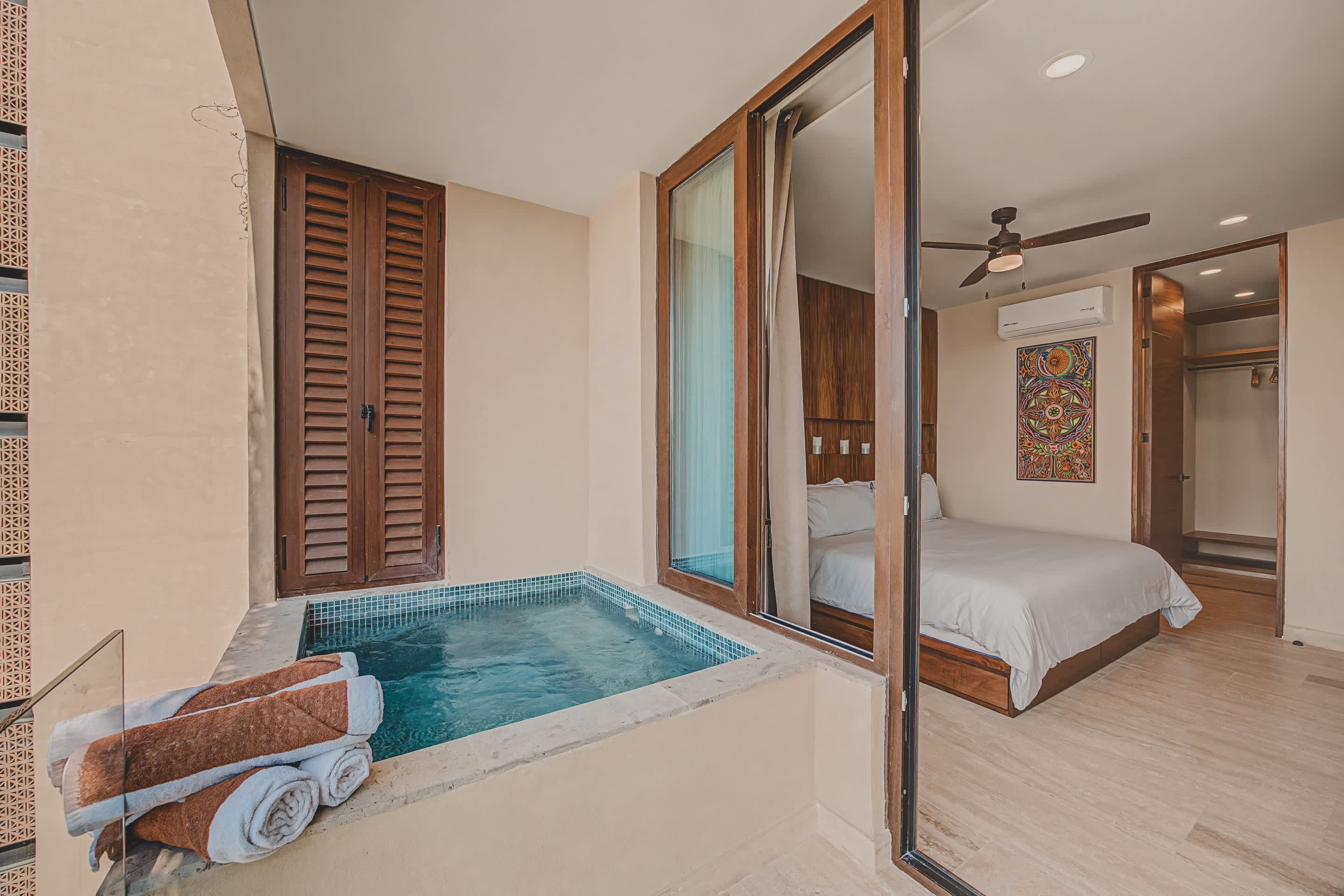 Modern bedroom with a white bed and wooden headboard, opening to a private balcony with a built-in tiled hot tub, stacked towels, and a view of the exterior.