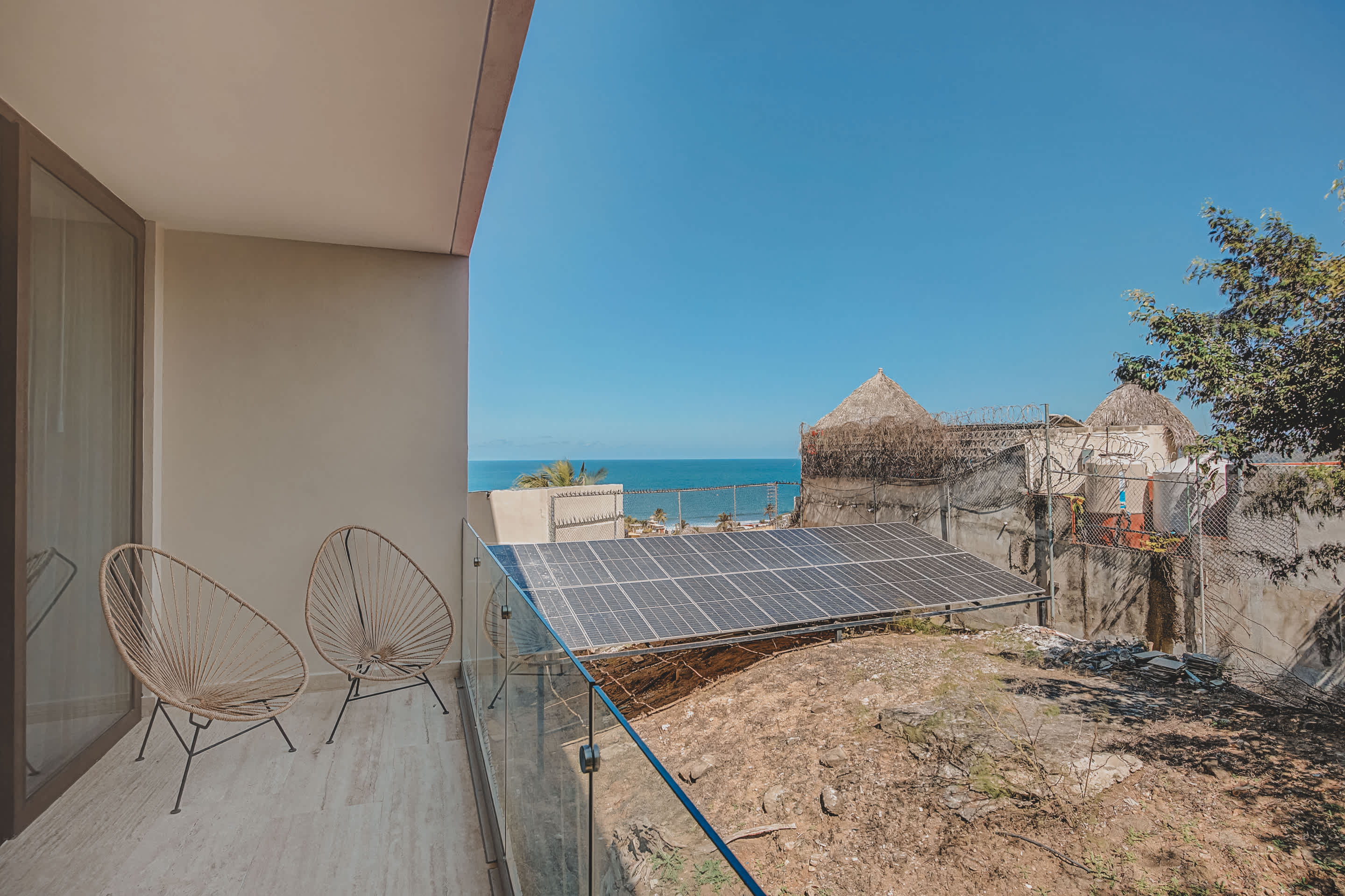 Modern balcony with light-colored tiled flooring, a clear glass railing, and two woven chairs, offering a view of the ocean, solar panels on a dirt patch, and distant buildings under a clear blue sky.