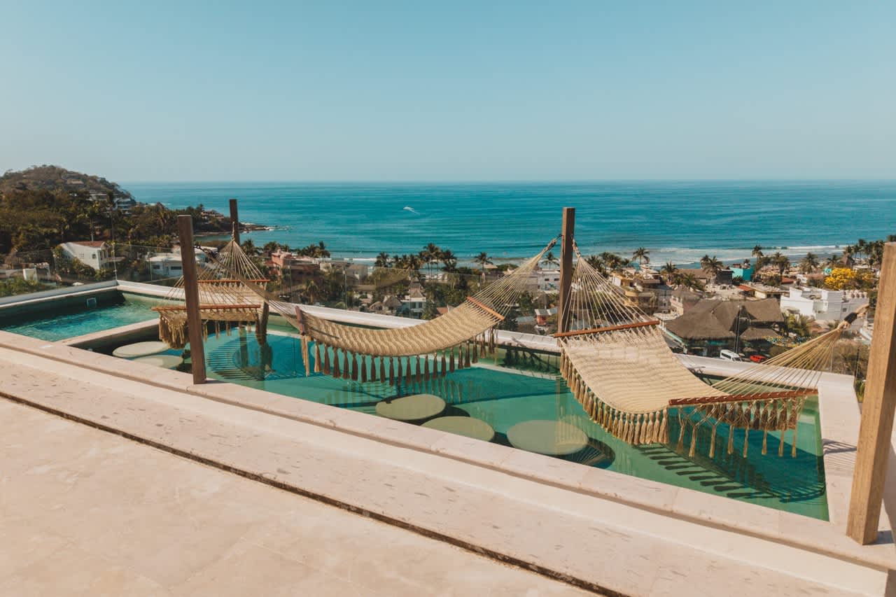 Elevated rooftop pool featuring two large hammocks suspended over the water, offering a panoramic view of the coastal town and the expansive ocean under a clear sky.
