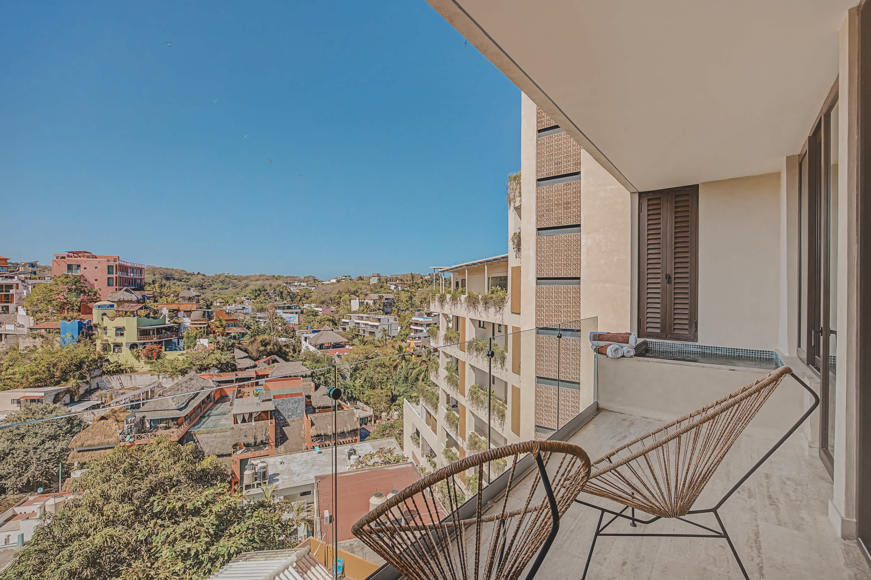 A spacious balcony offers panoramic views of a vibrant hillside town with diverse architecture and lush greenery, extending to the horizon under a clear blue sky. Two woven chairs provide seating, and a small built-in hot tub is visible on the right.