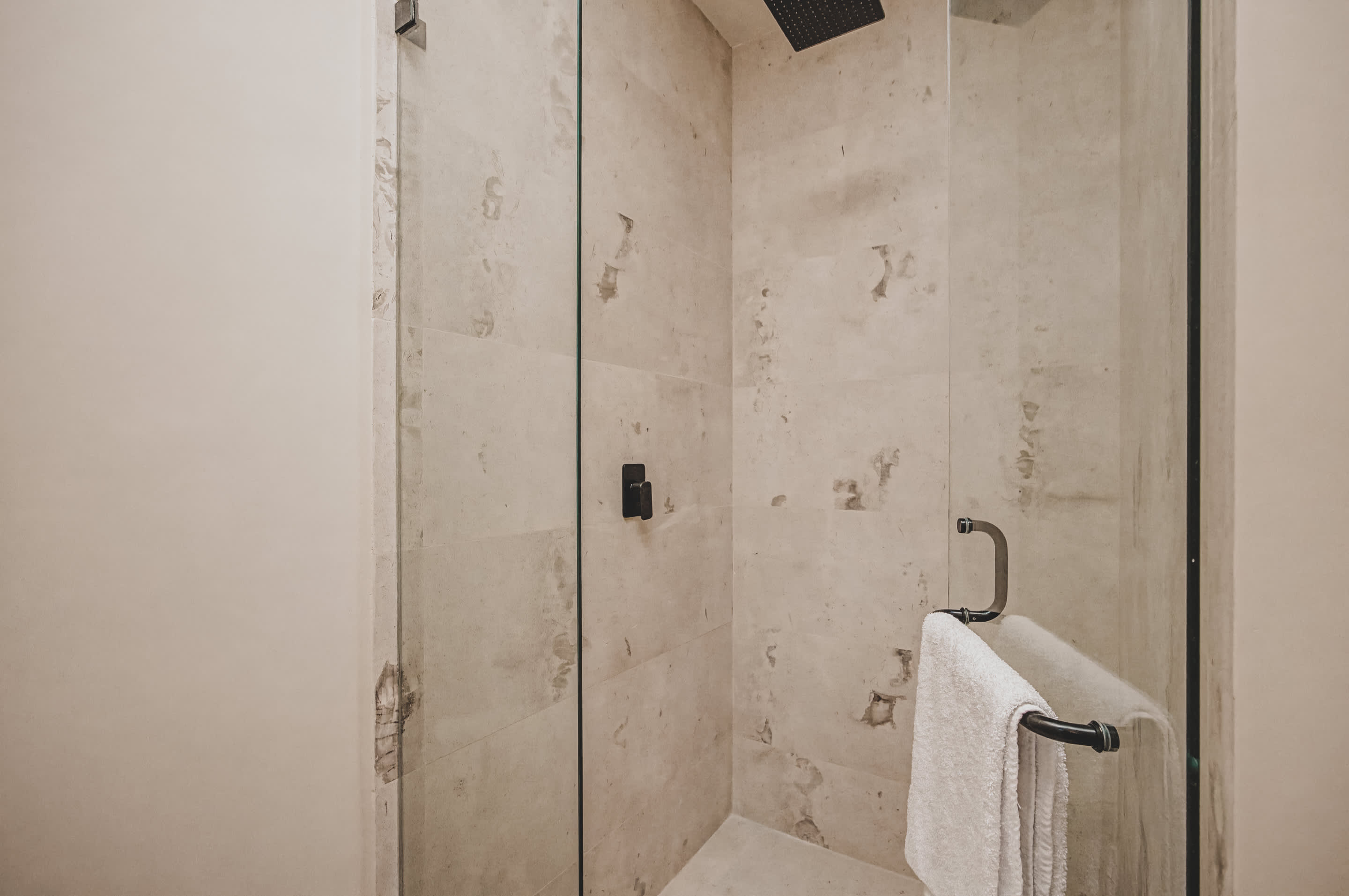 Spacious walk-in shower with light-colored stone-look tiles, a glass enclosure, and a dark rain shower head, featuring a towel hanging on the glass door handle.