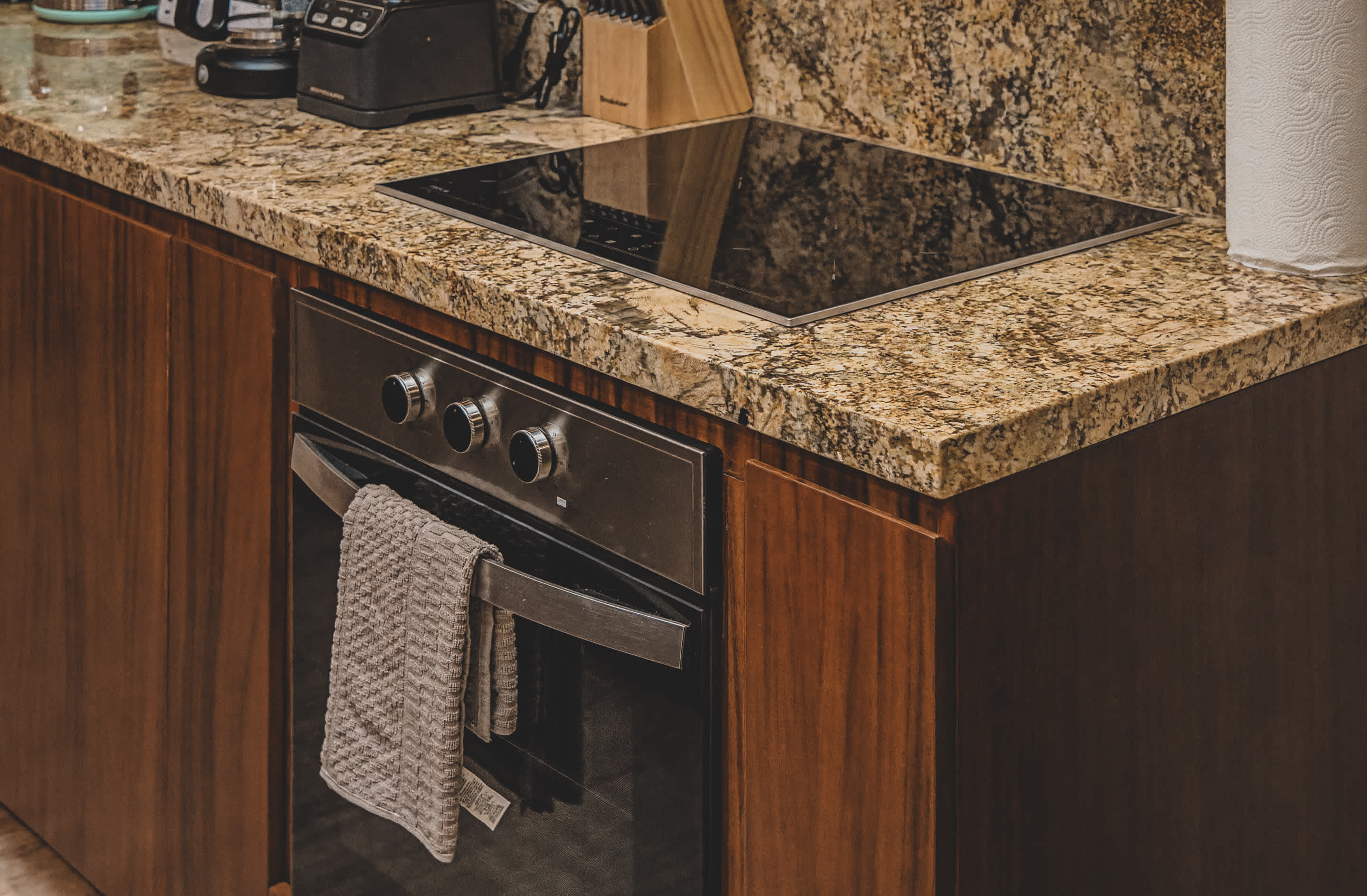 View of a kitchen area featuring a black induction cooktop and a stainless steel oven with a towel, all set within dark wood cabinets and a speckled granite countertop.