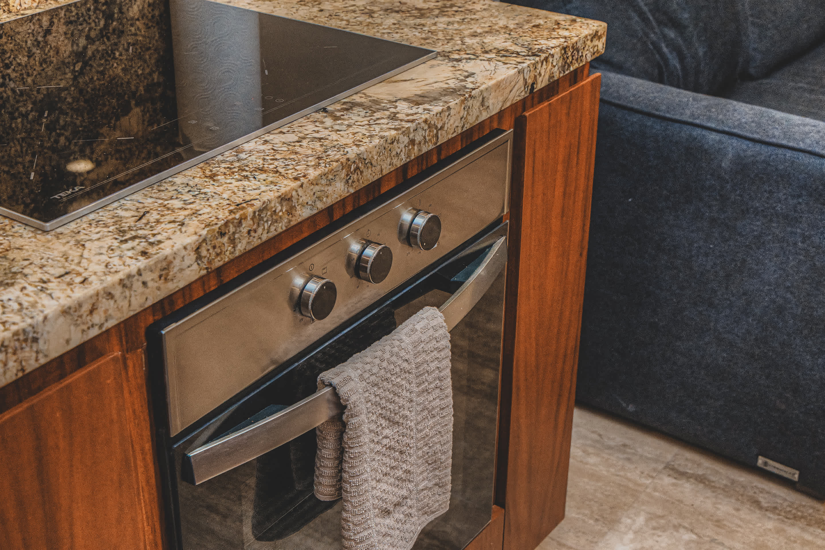 Stainless steel oven with control knobs and a towel hanging from its handle, integrated into dark wood cabinetry beneath a speckled granite countertop and black induction cooktop.