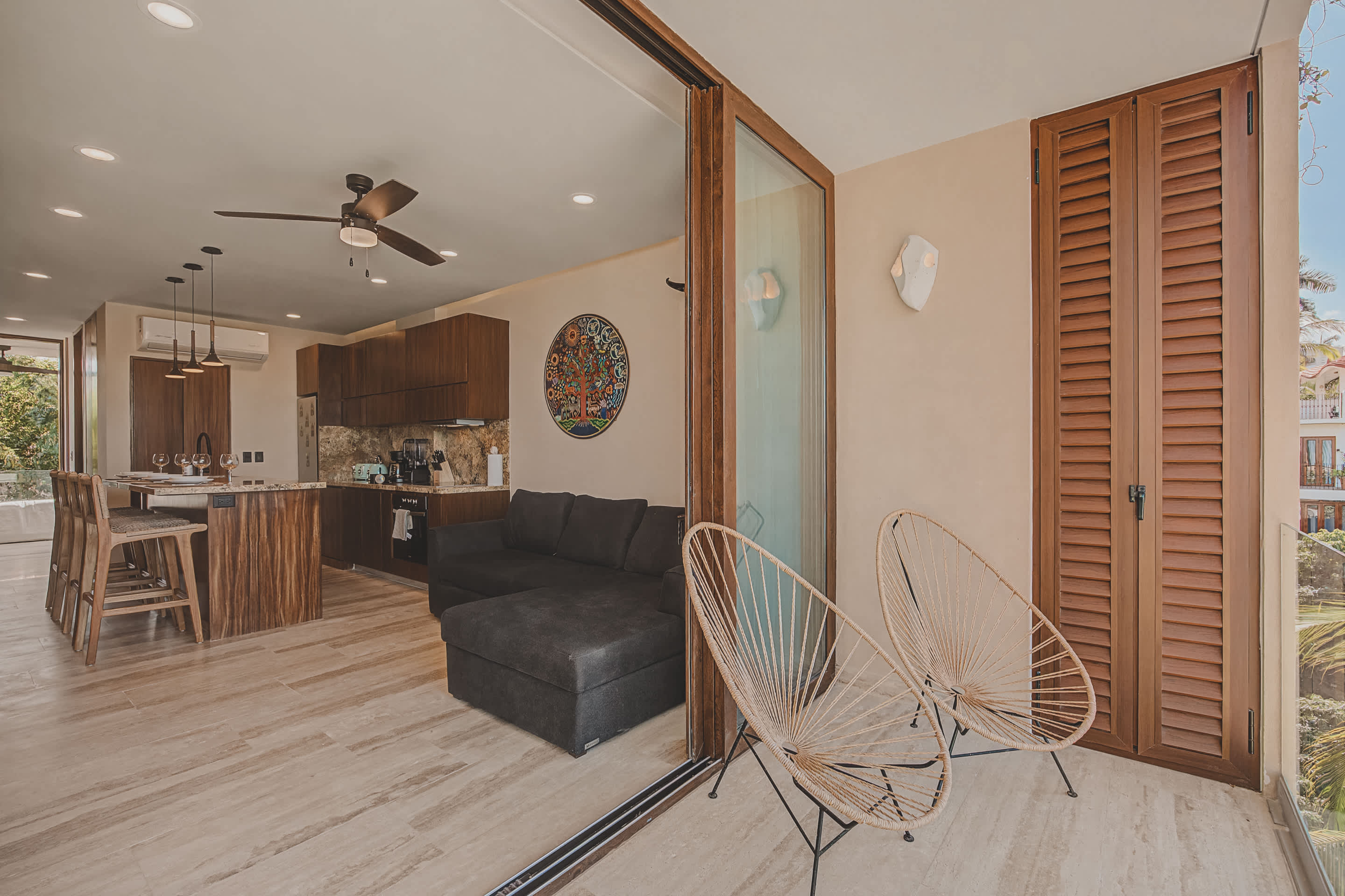 An open-concept living space features a modern kitchen with wood cabinetry and granite countertops, flowing into a living area with a dark sectional sofa. Large sliding glass doors open to a balcony with two woven chairs, offering an indoor-outdoor living experience.