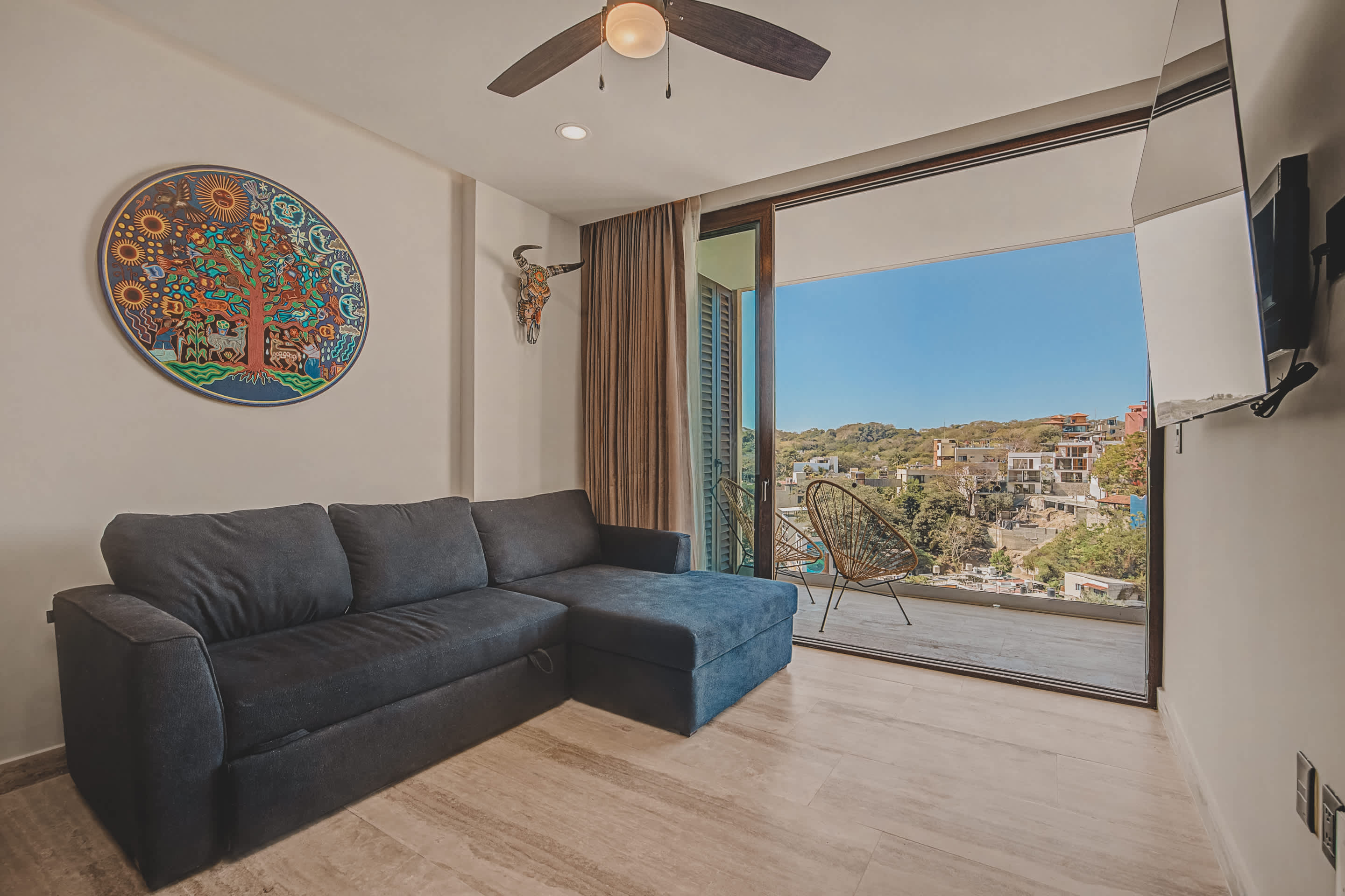 The cozy living room features a dark gray sectional sofa and a colorful circular wall art piece, with a ceiling fan overhead. Large sliding glass doors open to a balcony, revealing a vibrant cityscape and lush hills under a clear blue sky.