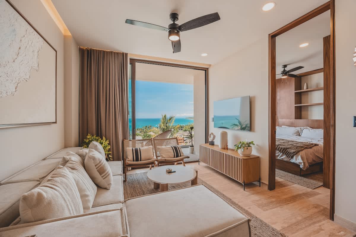 A bright living room features a large L-shaped sofa and a round coffee table, facing a wall-mounted TV. Sliding glass doors open to a balcony with an expansive ocean view, connecting to a bedroom on the right.