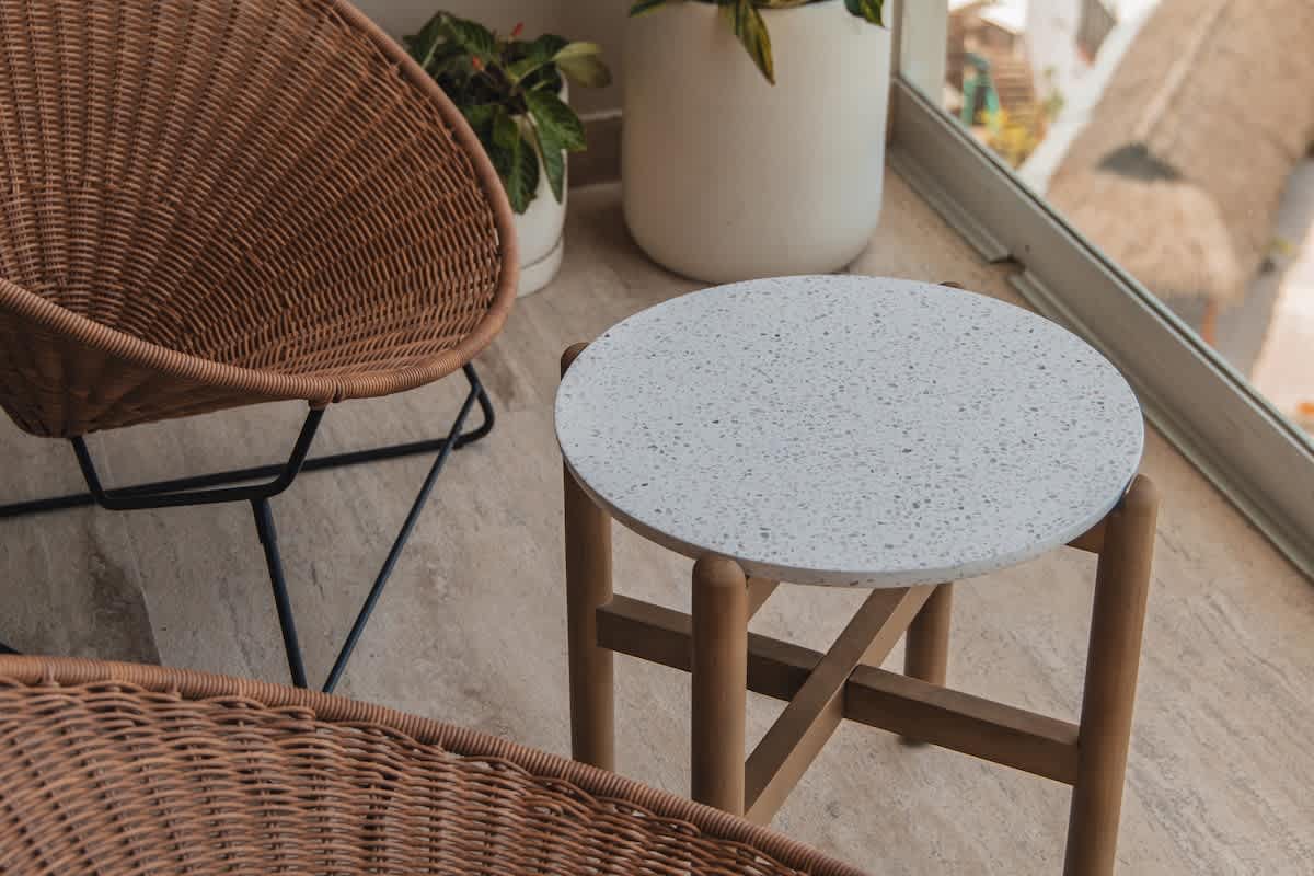 A round white terrazzo-top table with light wooden legs is positioned next to a woven wicker chair on a tiled balcony, with a glimpse of the ocean in the background.