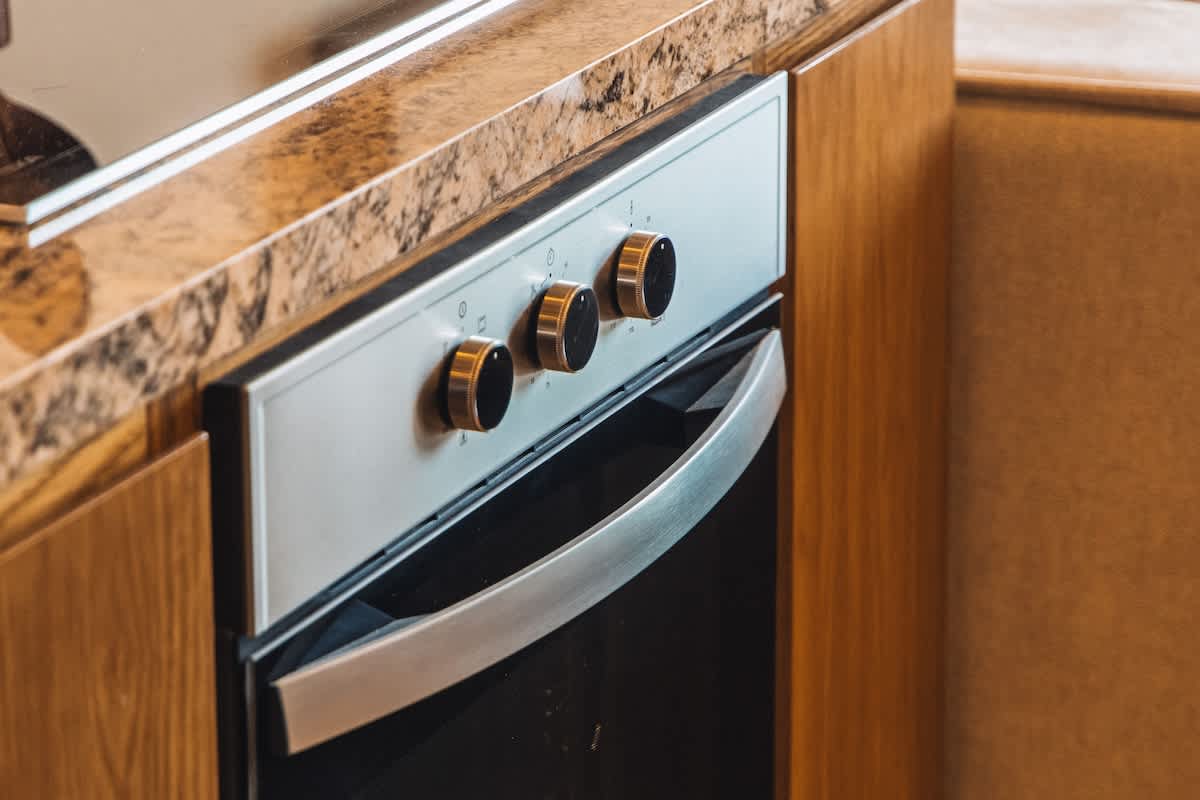 A close-up of a modern kitchen featuring a stainless steel built-in oven with black and gold knobs, set within light wooden cabinetry and topped with a speckled granite countertop.