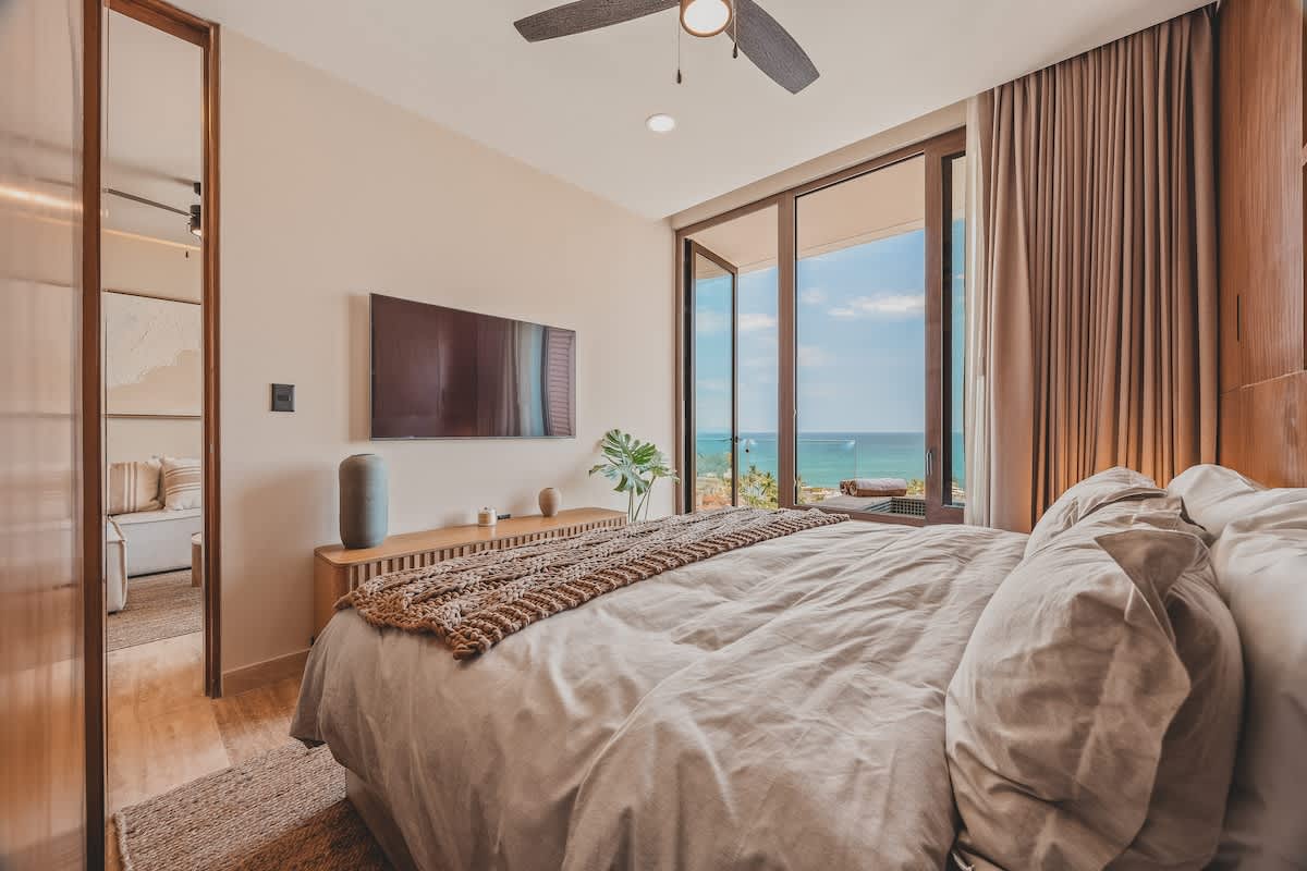 A bright bedroom with a large bed faces expansive sliding glass doors revealing a stunning ocean view. A wall-mounted TV and a wooden console table with a plant complete the modern, airy space.